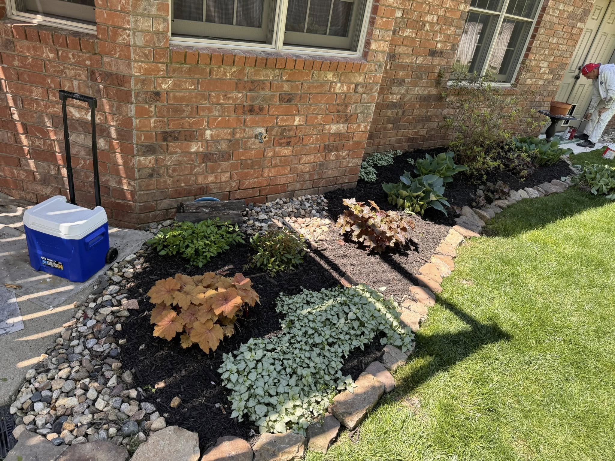 A person in white work clothes, red cap, using a trowel to work in a flower bed next to a brick house, with a cooler and gardening tools nearby.