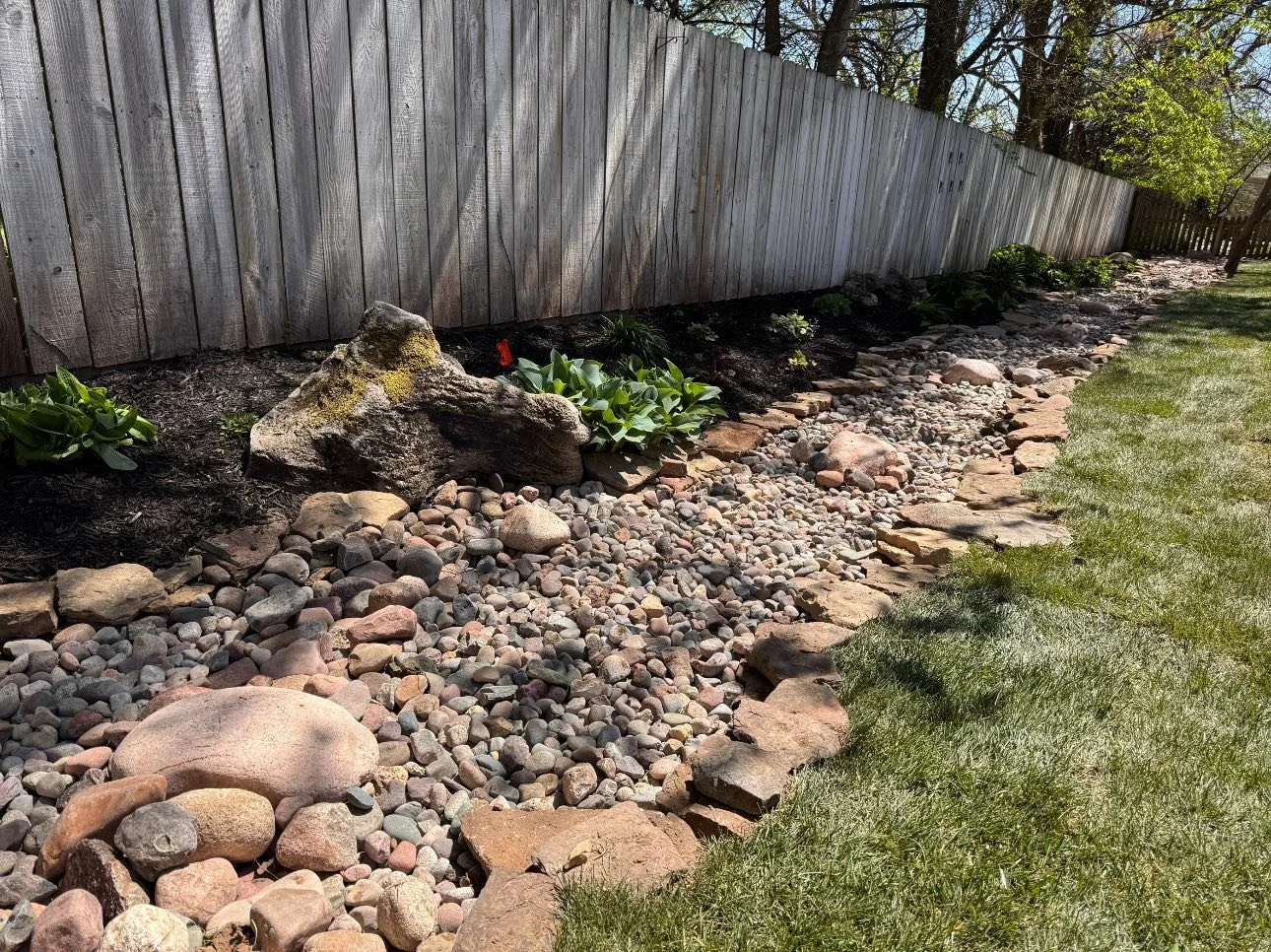 A garden bed with rocks, plants, and a weathered wooden fence in a backyard