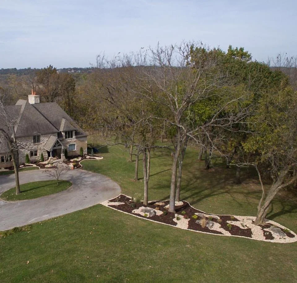 Residential yard with landscaped garden beds, leafless trees, and a house with a driveway in the foreground.