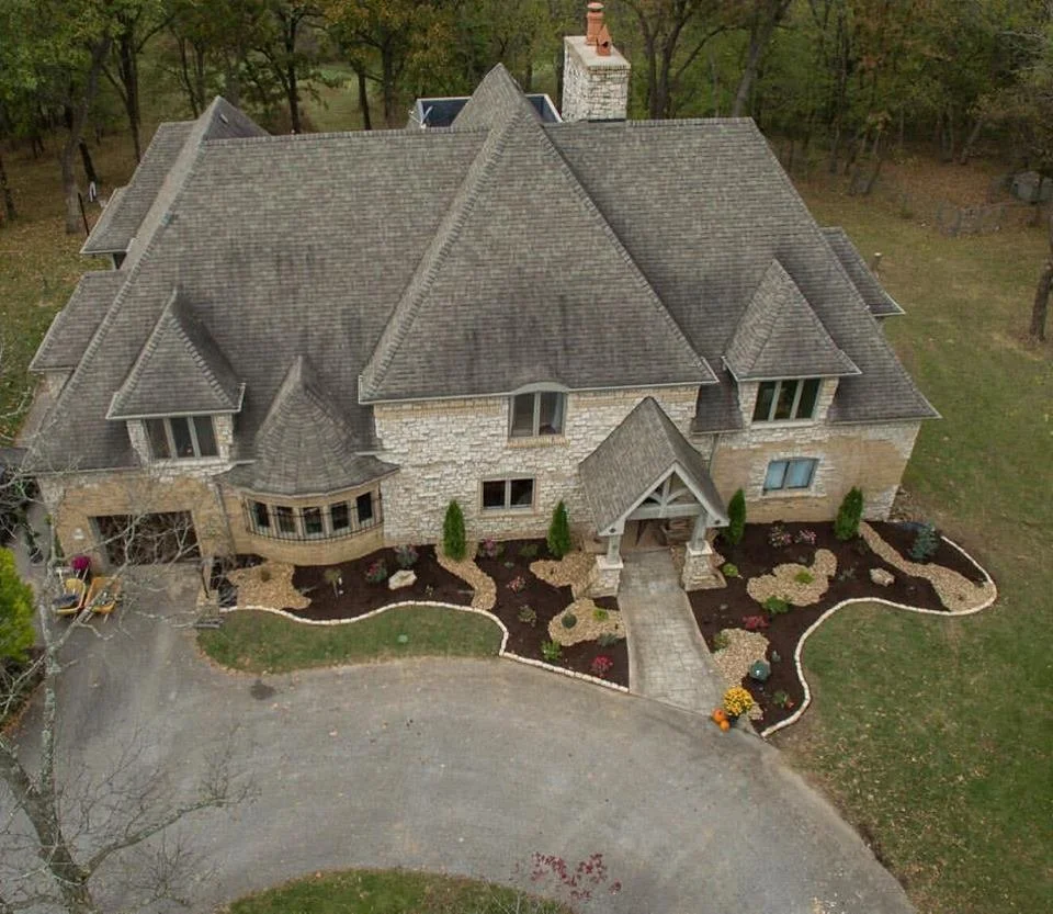 Aerial view of large house with stone exterior and steep roof, surrounded by landscaped yard with mulch and plants, located in a wooded area.