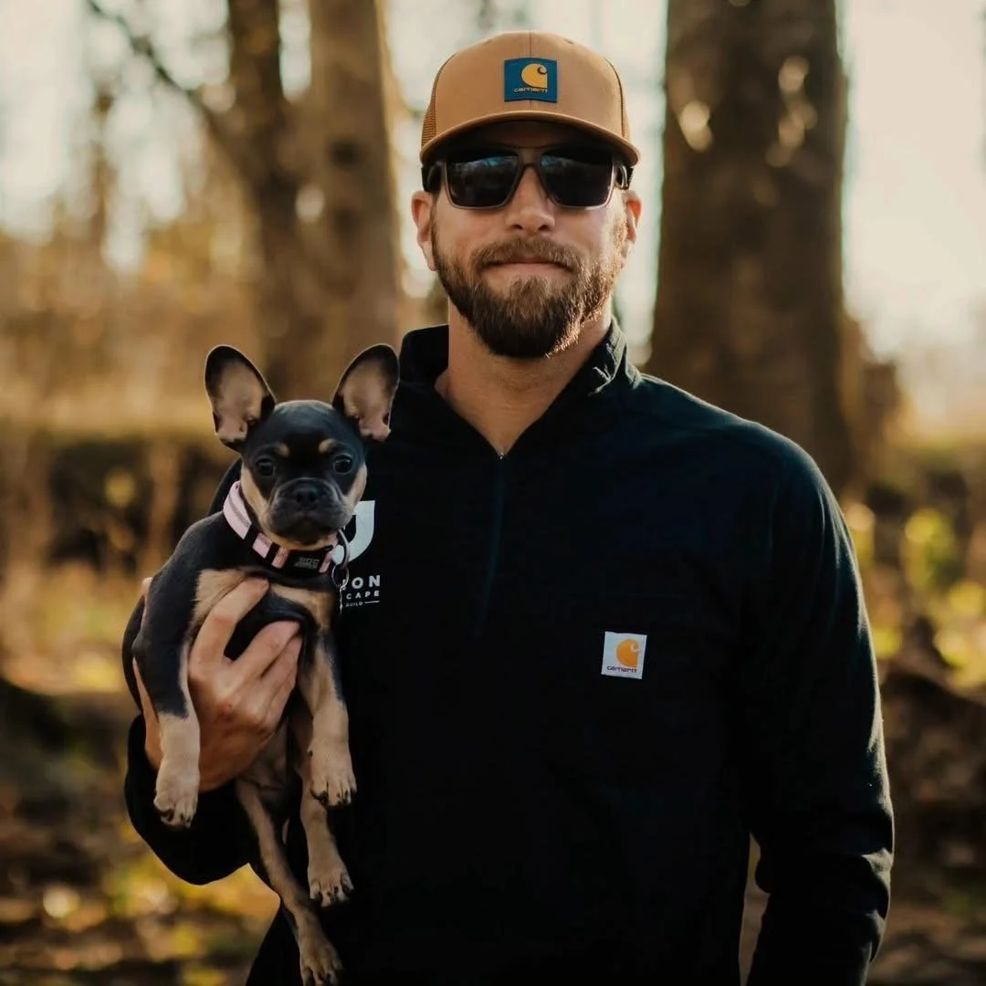 A man wearing sunglasses, a tan Carhartt cap, and a black jacket is holding a small black and tan Chihuahua with large ears outdoors in a wooded area.