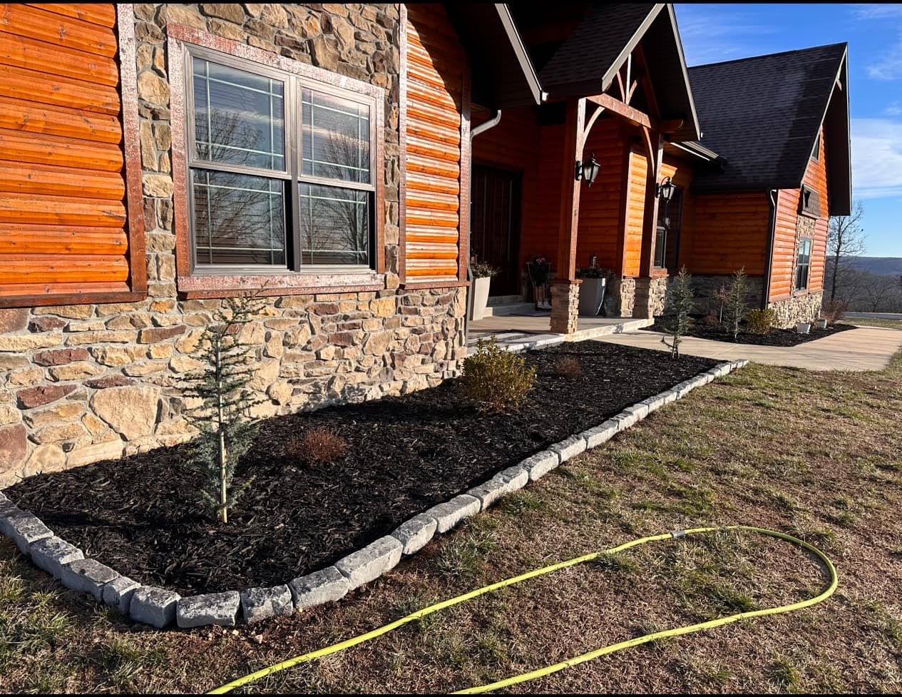 A newly landscaped front yard of a house with a mulched flower bed edged with stones, small trees, and a concrete walkway. The house has a stone and wood exterior, large windows, and a porch with two lantern-style lights.