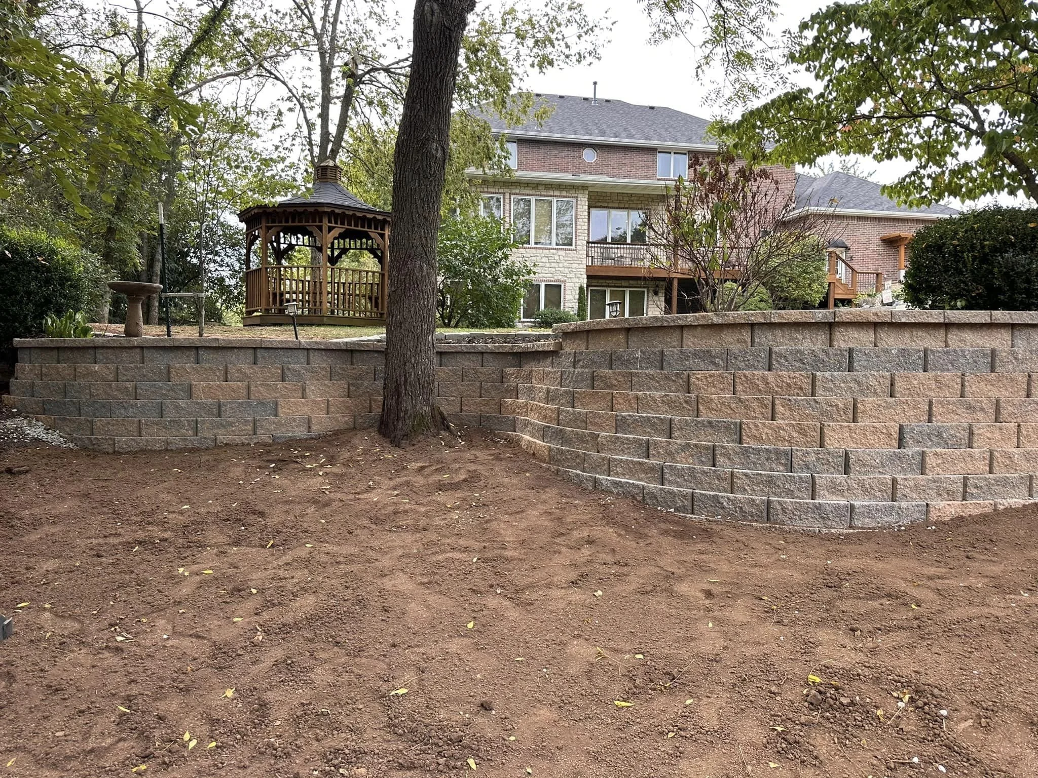 A backyard with brown dirt and a stone retaining wall, with a large tree, a gazebo, and a brick house with several windows and outdoor decks in the background.