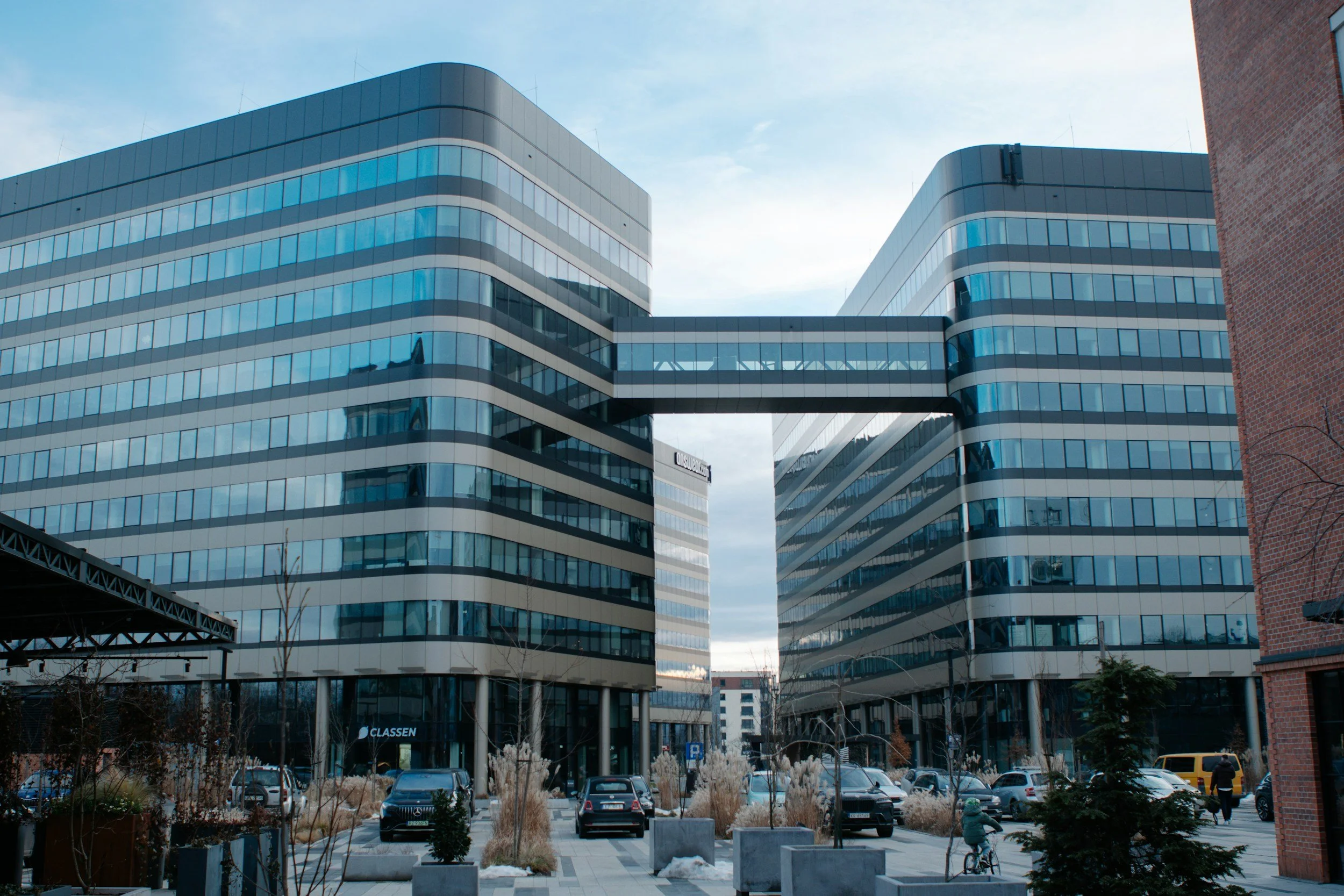Modern glass office buildings connected by a skybridge with a parking lot and trees in the foreground.