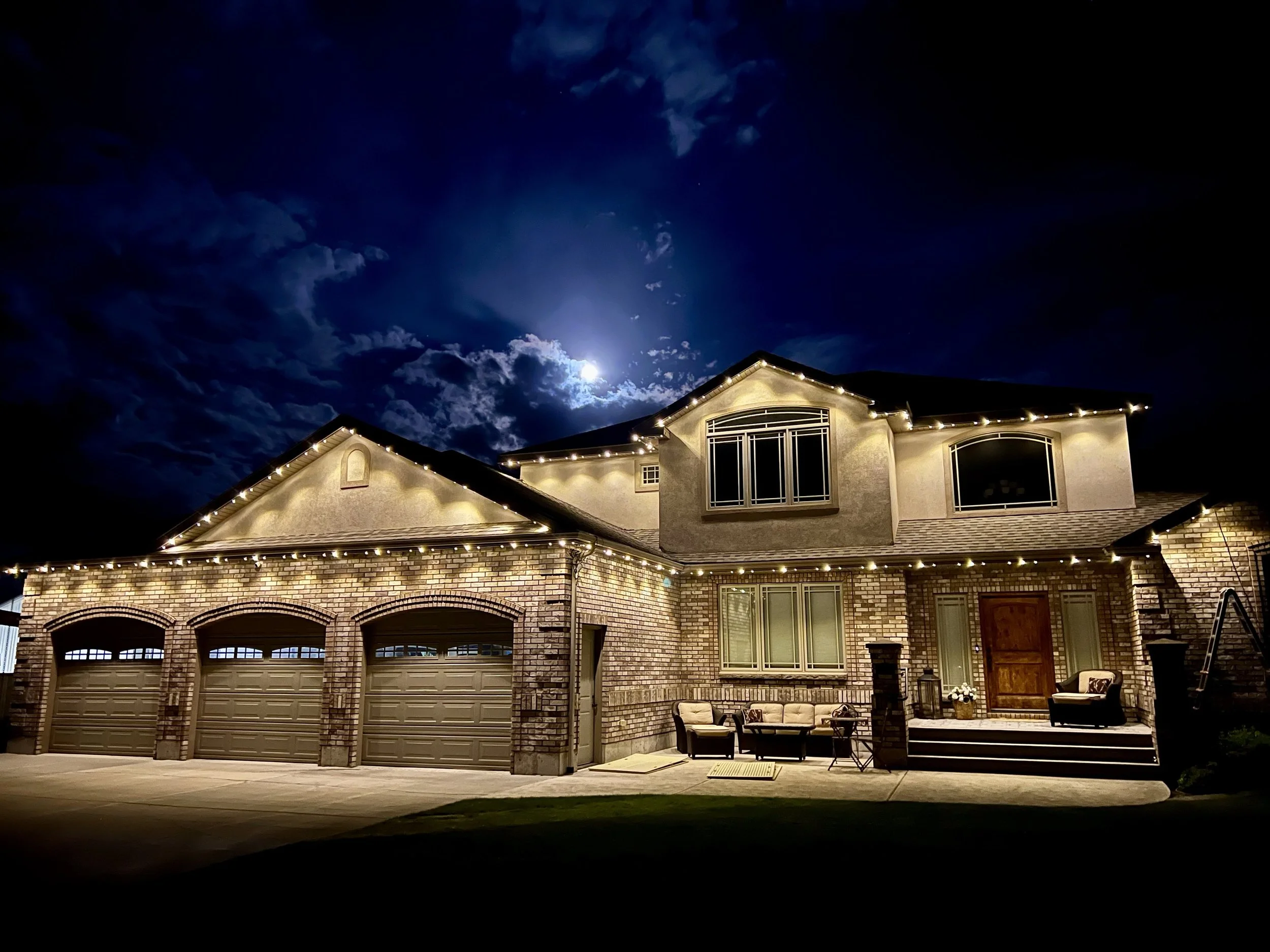 Nighttime view of a suburban house with string lights along the roofline, brick exterior, multiple garage doors, and a small outdoor seating area, under a partly cloudy sky with the moon shining.