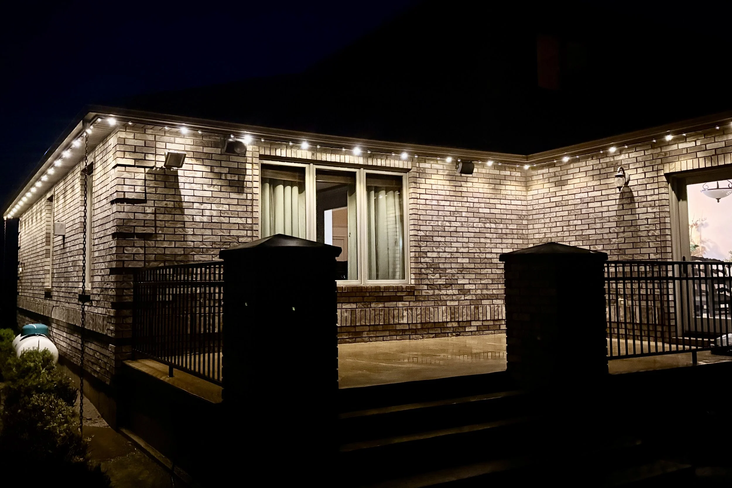 Nighttime view of a brick house with string lights along the roofline, a sliding glass door with curtains, a porch with a railing, and outdoor lighting.