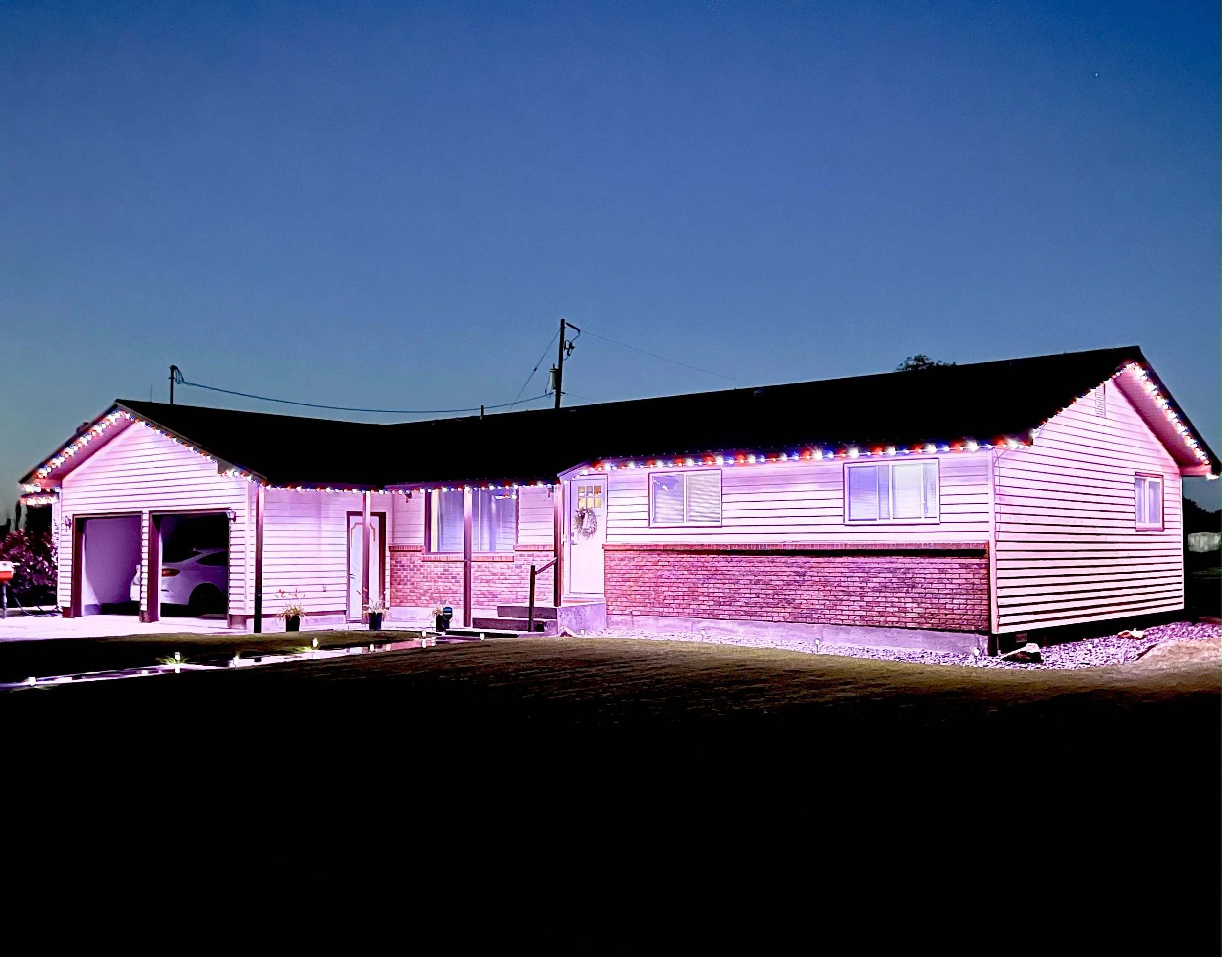 A house decorated with pink and purple Christmas lights on the roofline and around the windows, set against a dark evening sky.