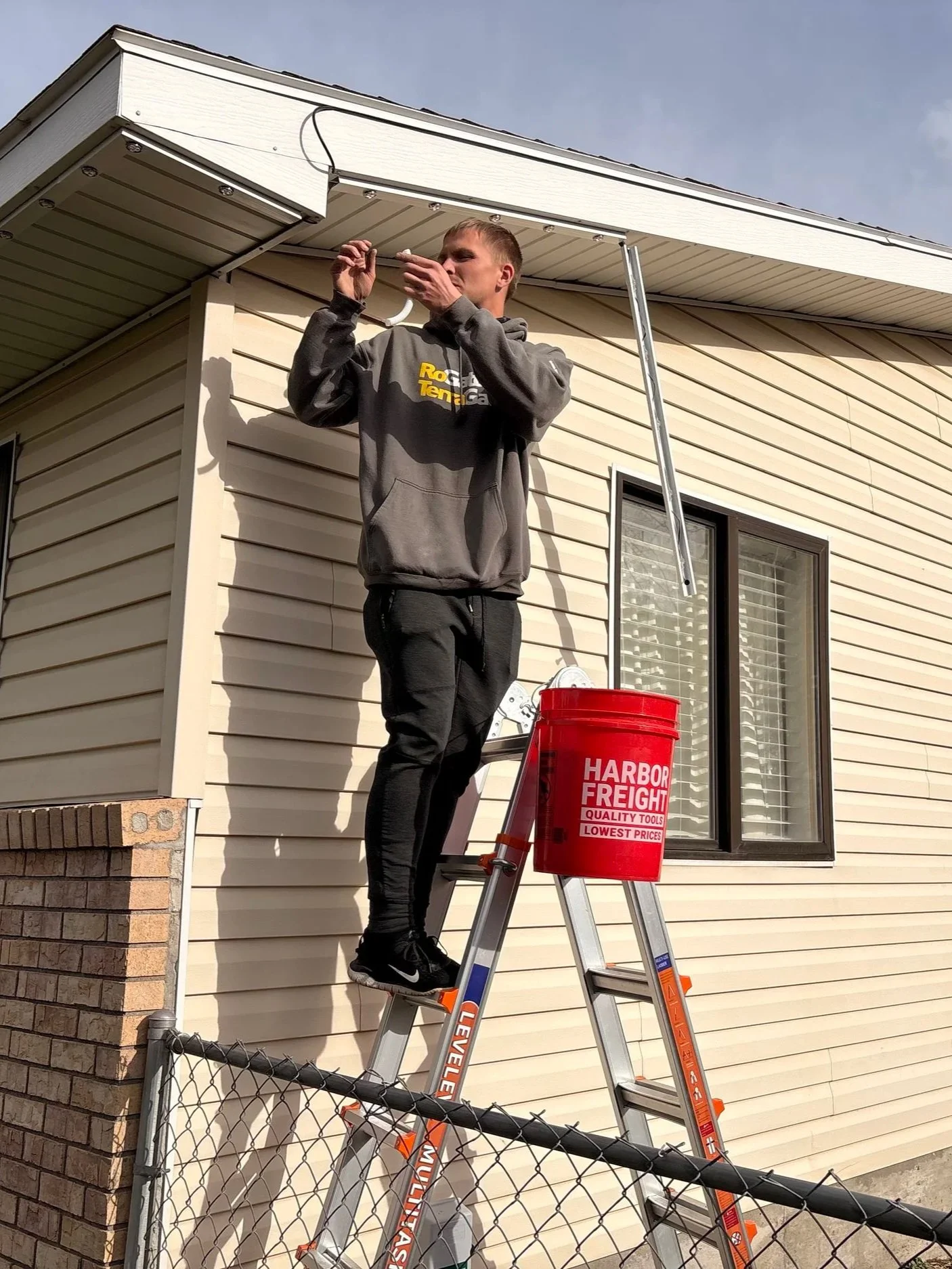 A young man standing on an extension ladder cleaning the gutter of a house. He is wearing a gray hoodie and black pants, and is holding a small tool in his right hand. The ladder is positioned against the house, which has light-colored siding and a window. There is a red bucket labeled 'Harbor Freight' on the ladder.