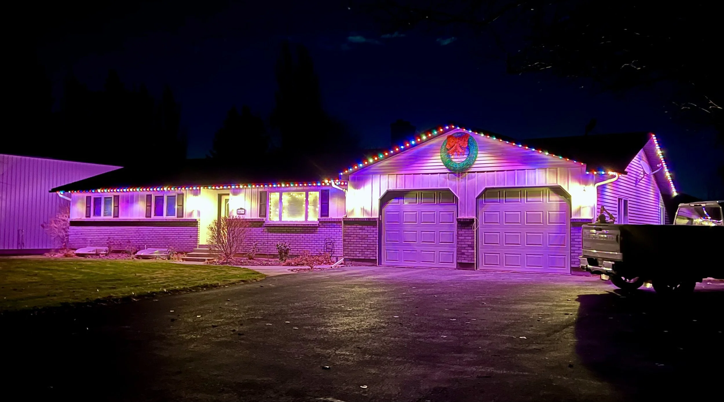 A house decorated with colorful Christmas lights at night. The house has a brick and siding exterior, and a garage with two doors. There is a festive wreath on the front of the house, and a Christmas figure is on the right side near the garage. The yard is grassy with some small plants, and a truck is parked on the driveway.