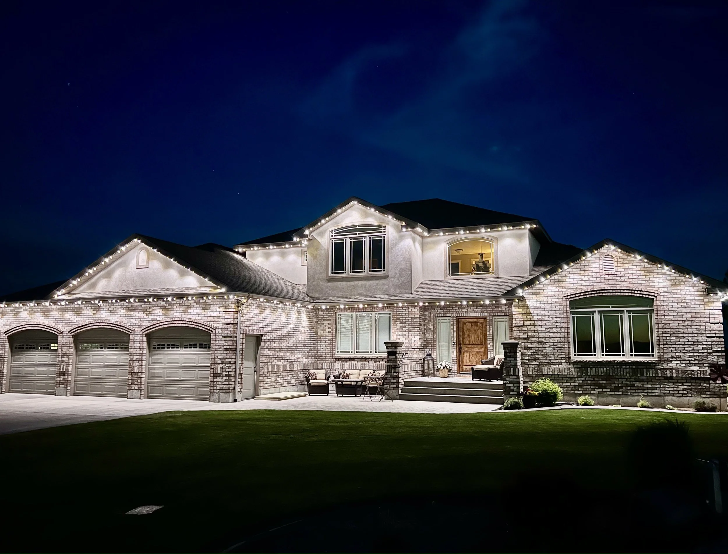 Nighttime view of a large house with a peaked roof, brick exterior, and multiple windows, decorated with string lights, with a front yard and patio area.