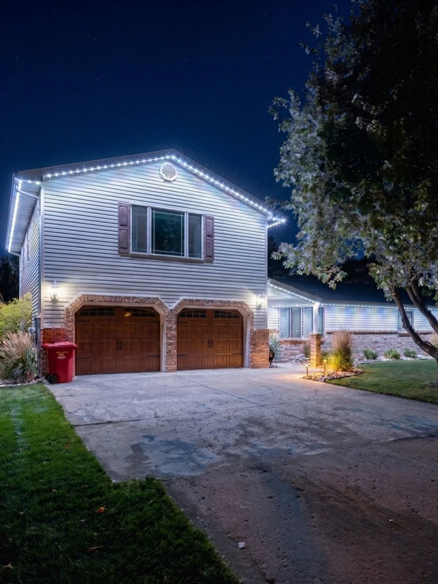 A house decorated with white string lights outlining the roof, with a two-car garage, a red trash bin, and a well-lit driveway at night.