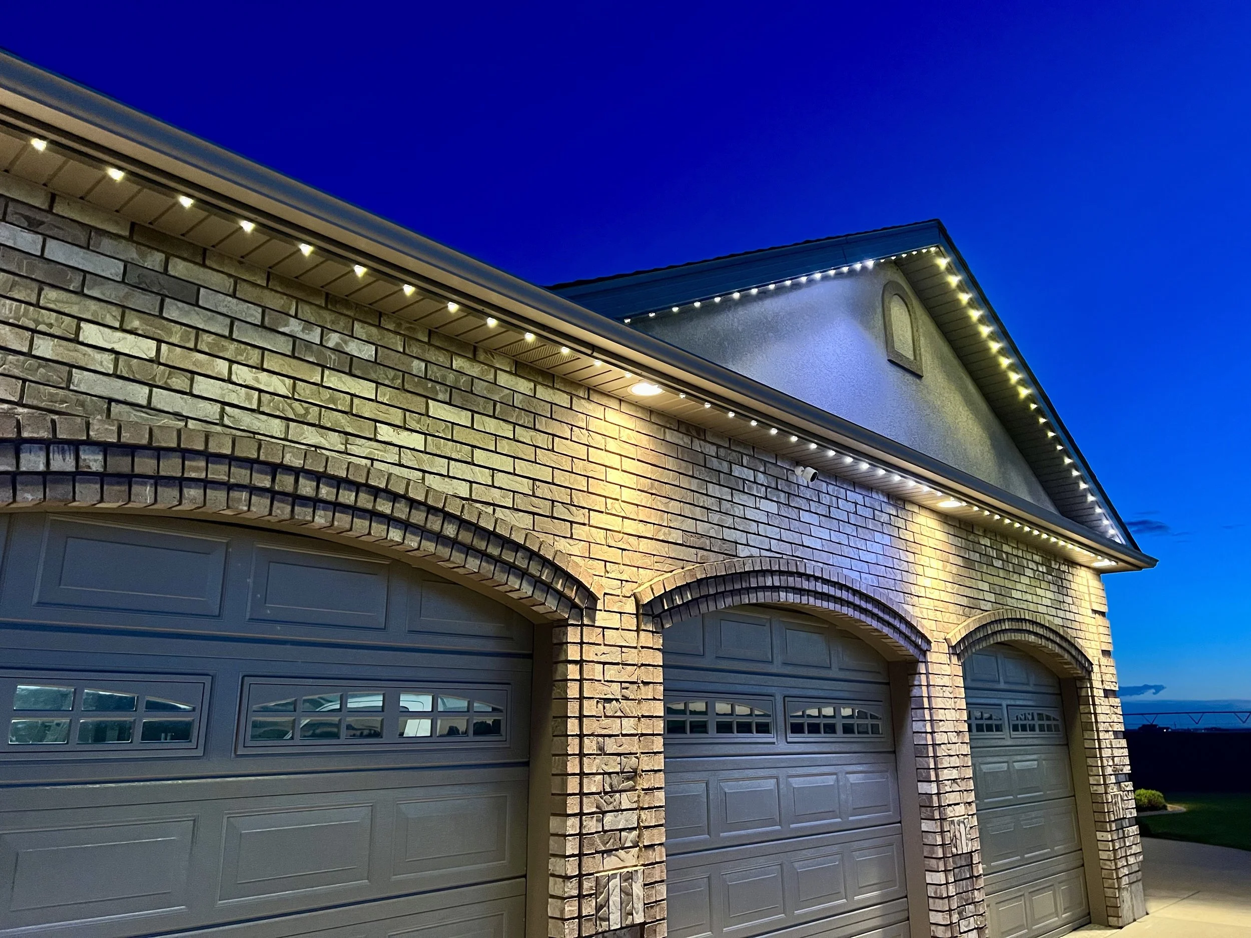 Two garage doors in a modern suburban house at dusk with exterior lighting and a brick wall facade.
