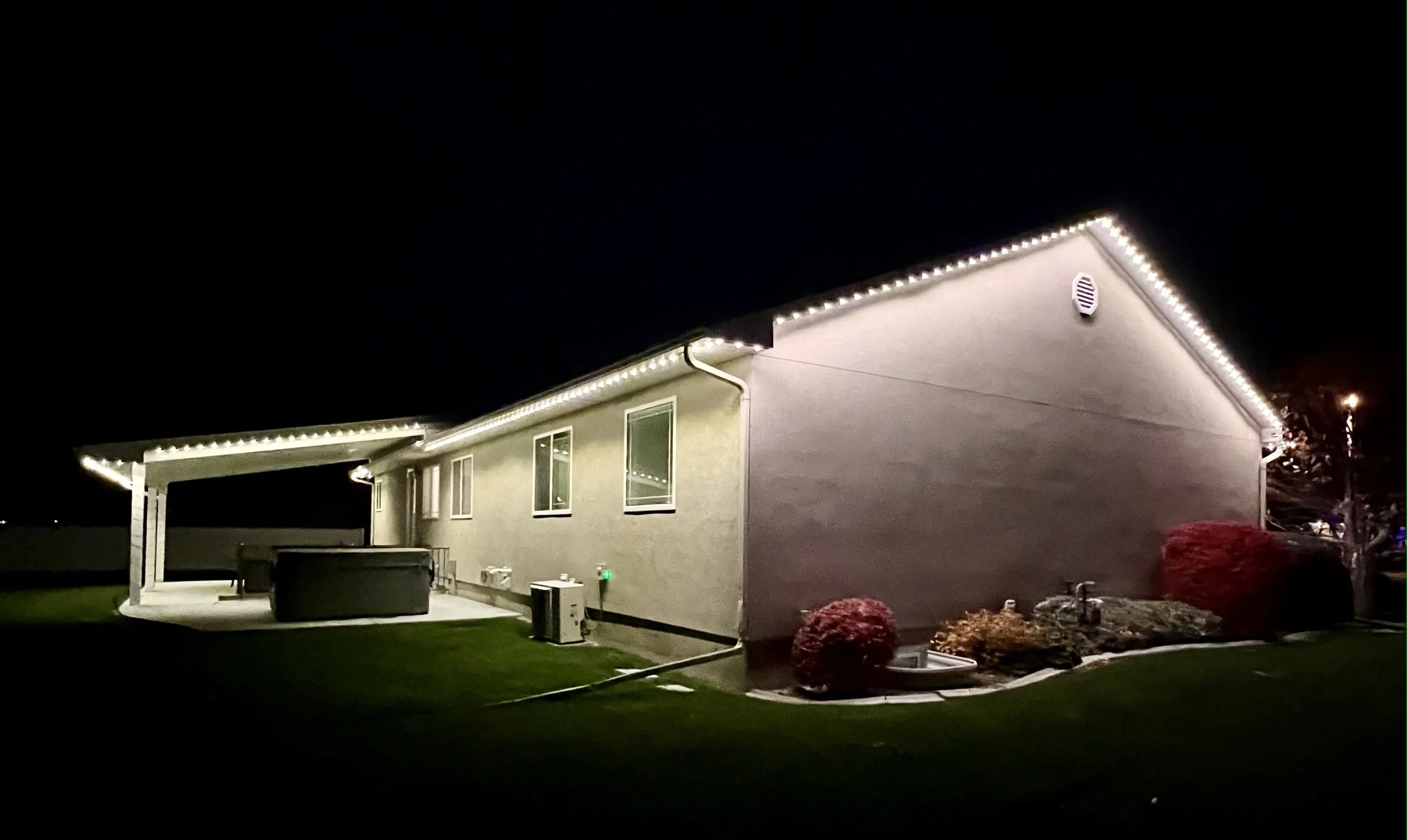 House at night with exterior lighting along roofline, a patio area with a covered structure, and landscaped yard with bushes and outdoor equipment.