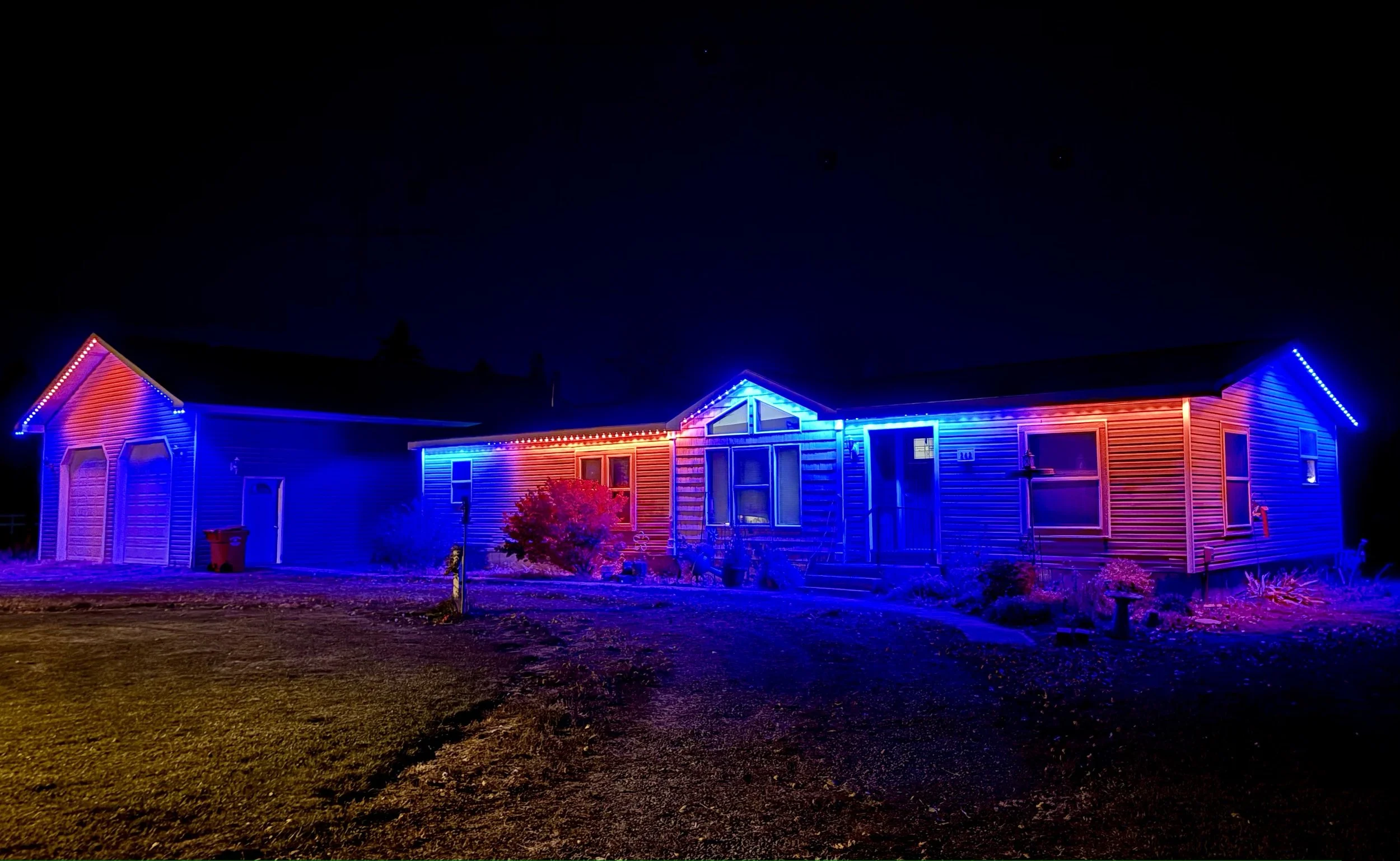 A house decorated with colorful Christmas lights featuring red, blue, and purple LED strip lights outlining the roof and windows, with a dark night sky above.