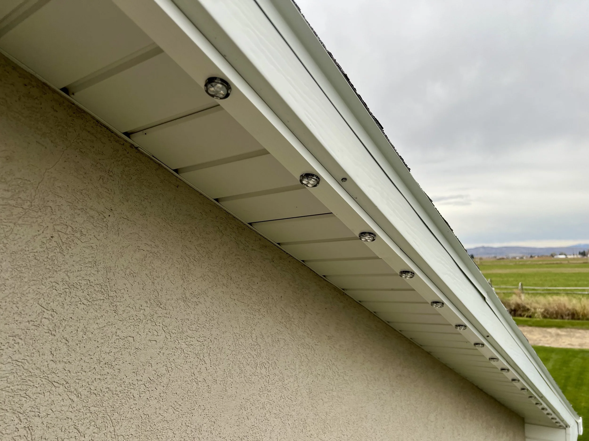 Close-up of the underside of a house's roof overhang with external lighting fixtures, beige textured wall, and a background of distant fields and mountains under a cloudy sky.