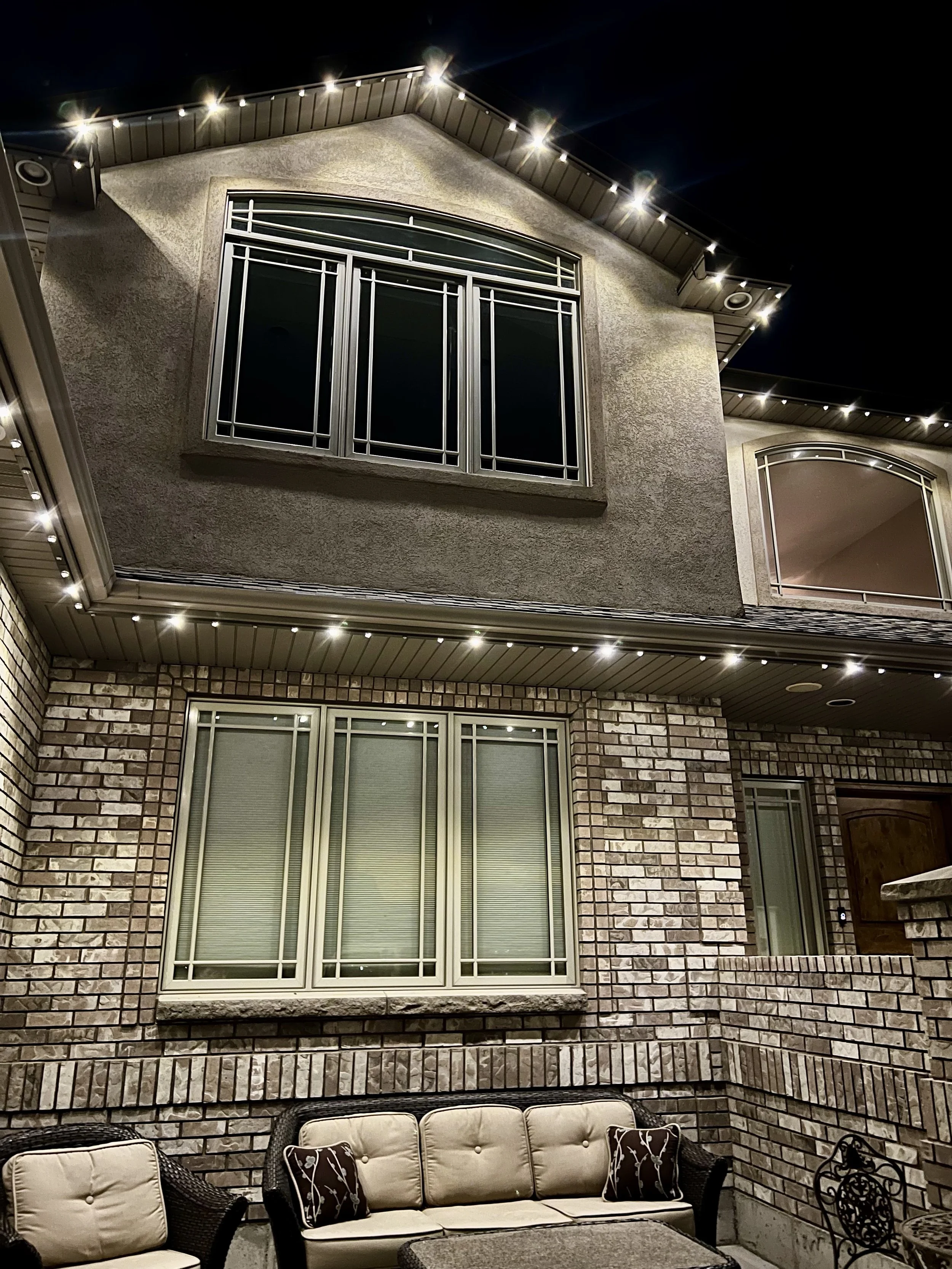 An outdoor patio area at night with a brick wall, a cushioned outdoor sofa with pillows, and large windows on the house. The house has a beige stucco exterior, metal window frames, and is decorated with string lights around the roof.