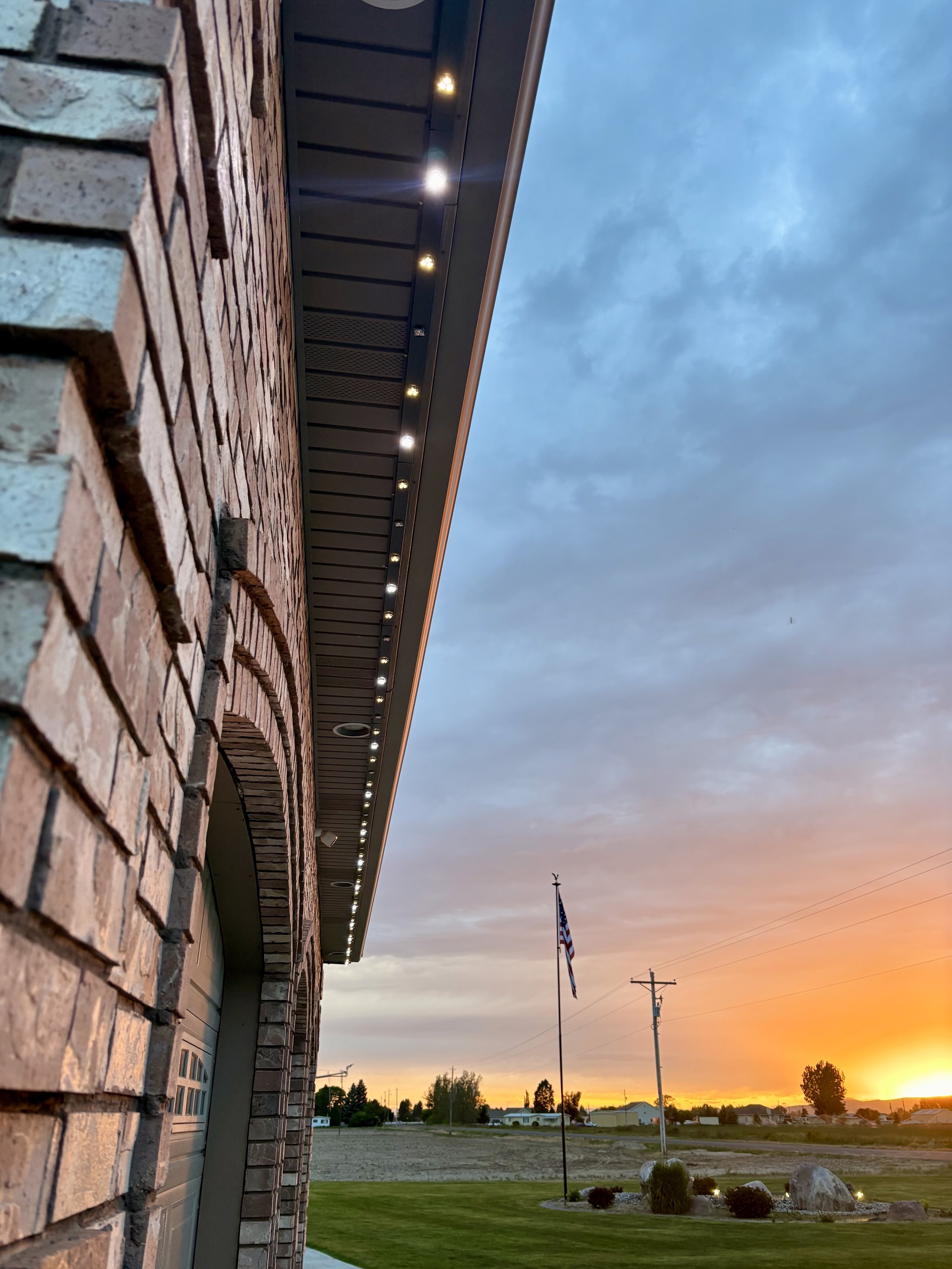 Side view of a brick building with exterior lighting under a dusk sky, with an American flag on a pole and a landscape with trees and rocks in the background.