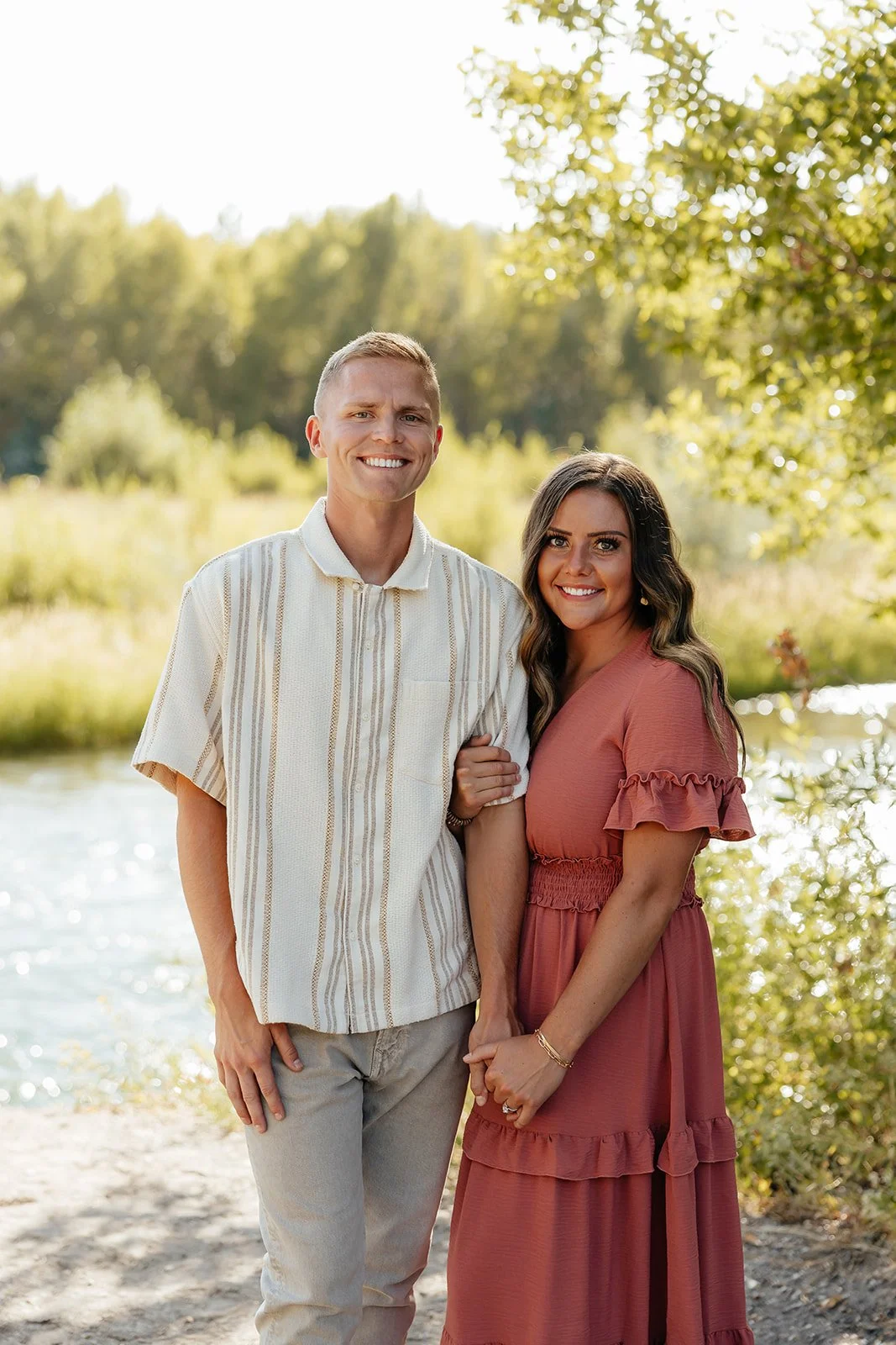 A smiling man and woman standing outdoors near a water body, with green trees in the background during daytime.