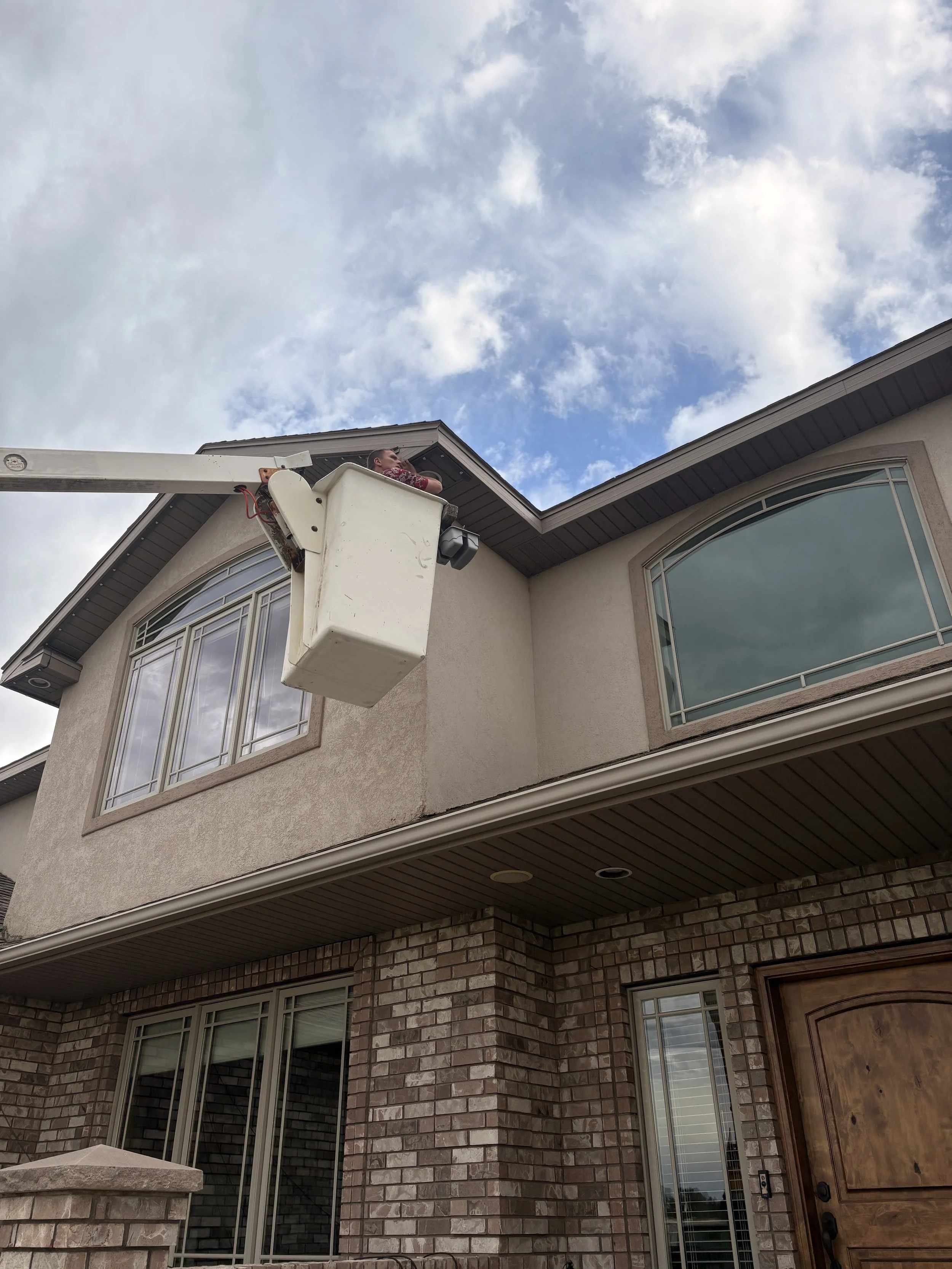 A person working on a house roof using a bucket truck, with a blue sky and scattered clouds overhead.
