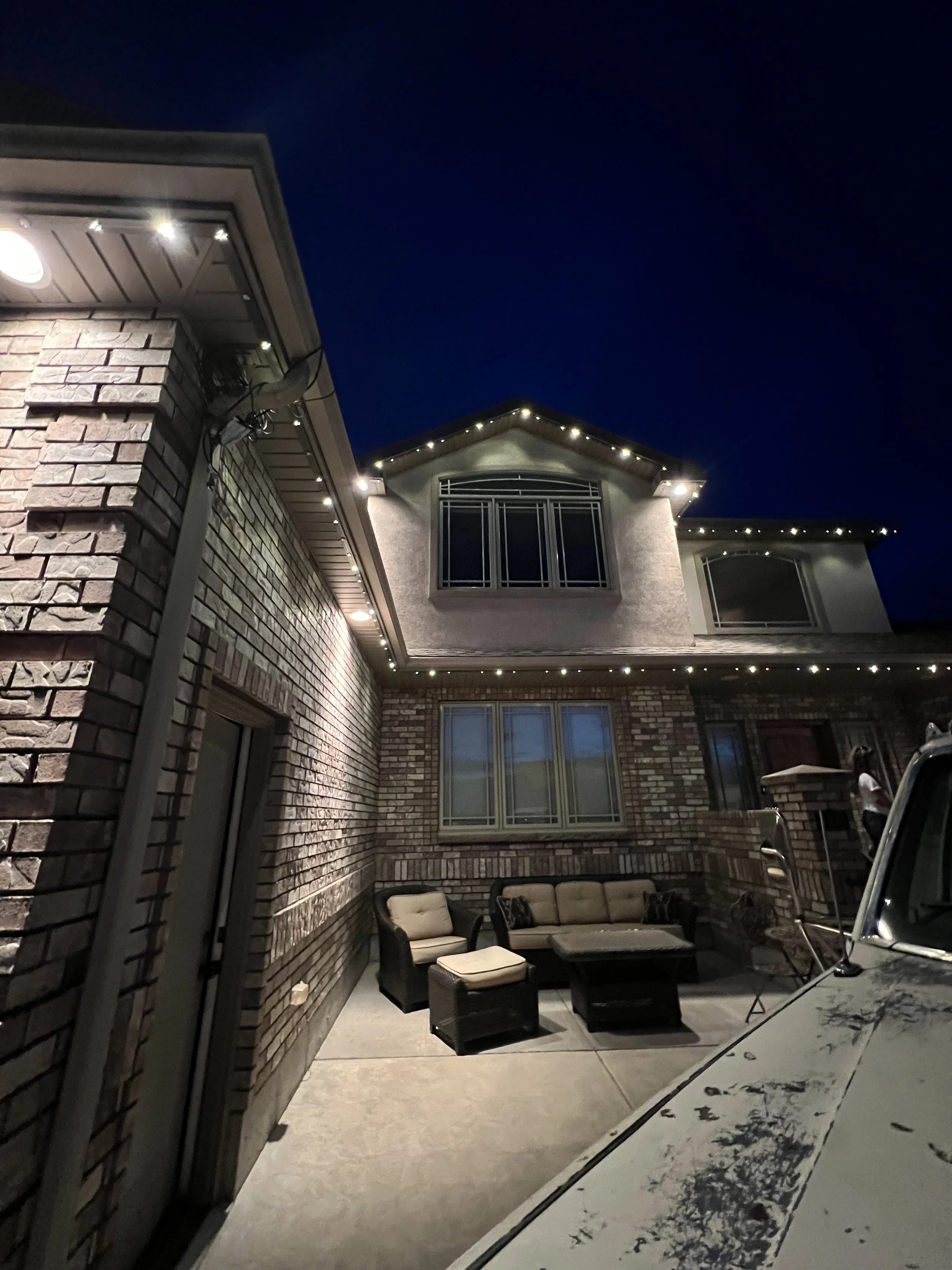 Nighttime scene of a brick house exterior with string lights, patio furniture including a sofa, chairs, and a table on a concrete patio, and a large window with a balcony. Part of a car is visible in the bottom right corner.