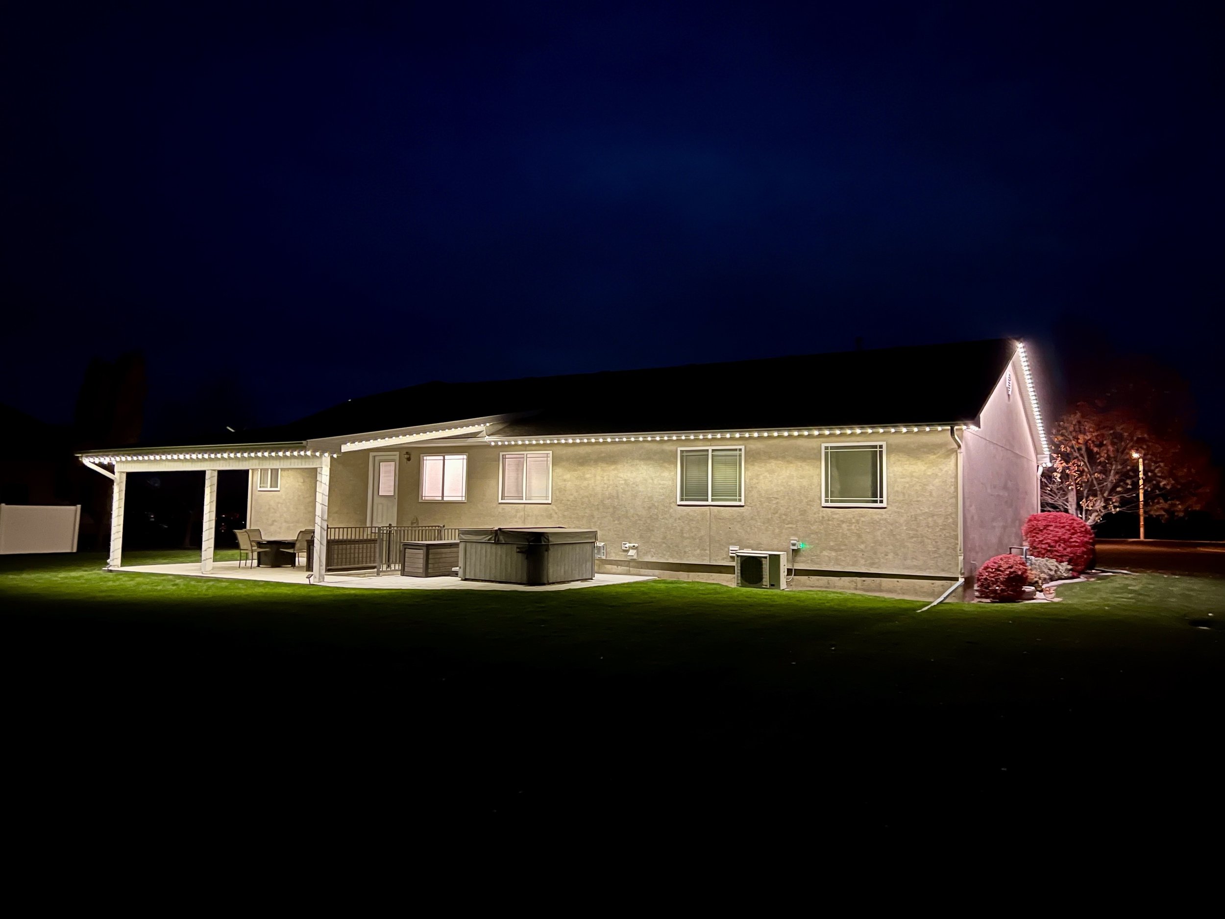 A house at night with string lights outlining the roof, a covered patio area with outdoor furniture, and a well-maintained lawn with some bushes and trees.