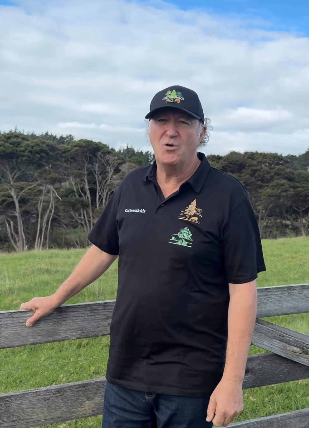 A man outdoors standing by a wooden fence, wearing a black cap and a black polo shirt with logos of trees and the word 'Carbonfields', on a cloudy day with green fields and trees in the background.