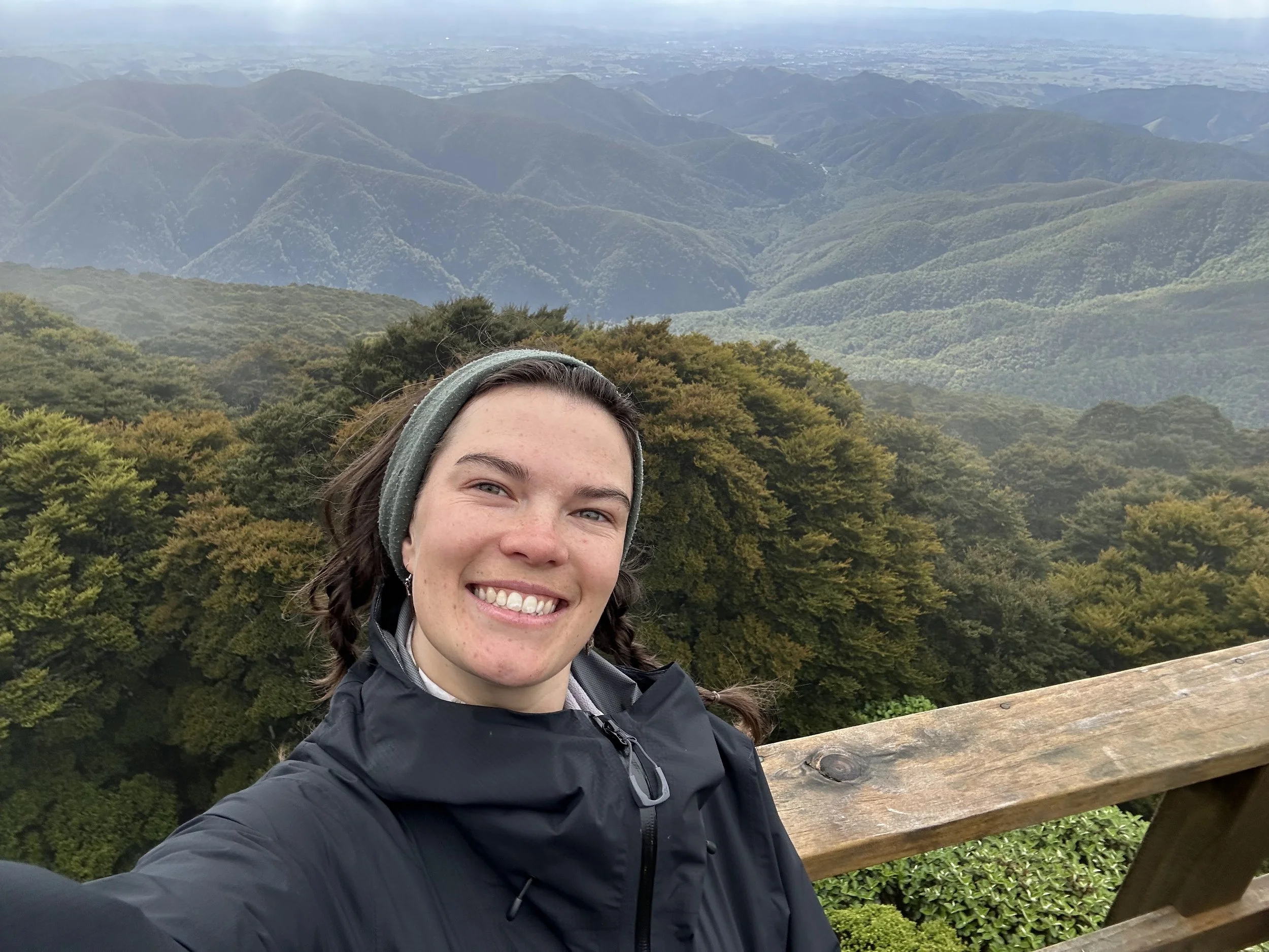 A smiling woman with brown hair and a headband taking a selfie on a mountain trail with lush green trees and mountain valleys in the background.