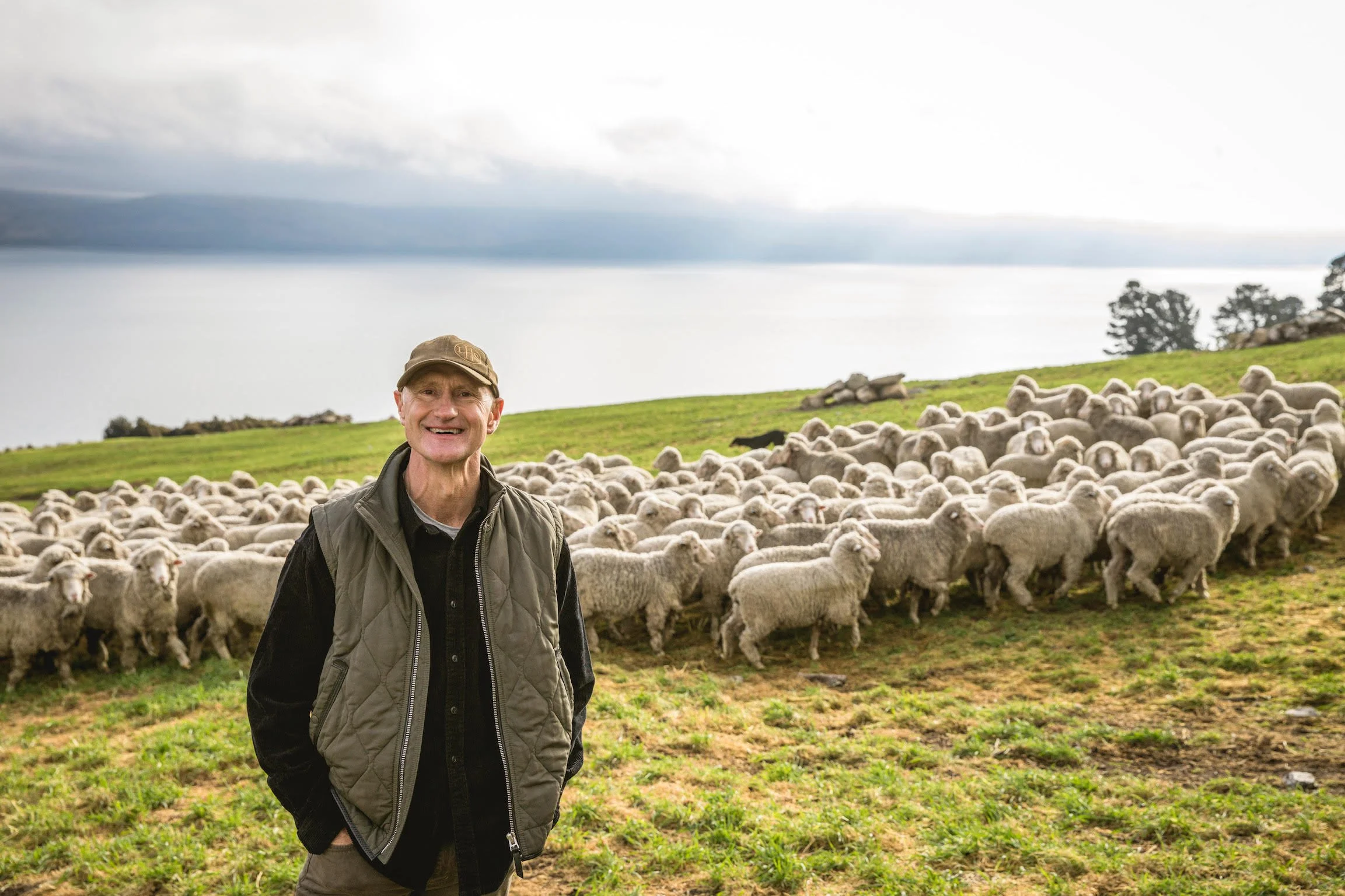 A man smiling in front of a flock of sheep on a grassy hillside with trees and a cloudy sky in the background.