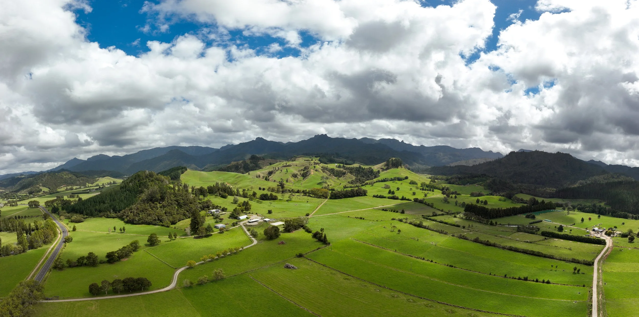 Scenic landscape of green rolling hills, scattered farms, and distant mountains under a partly cloudy sky.