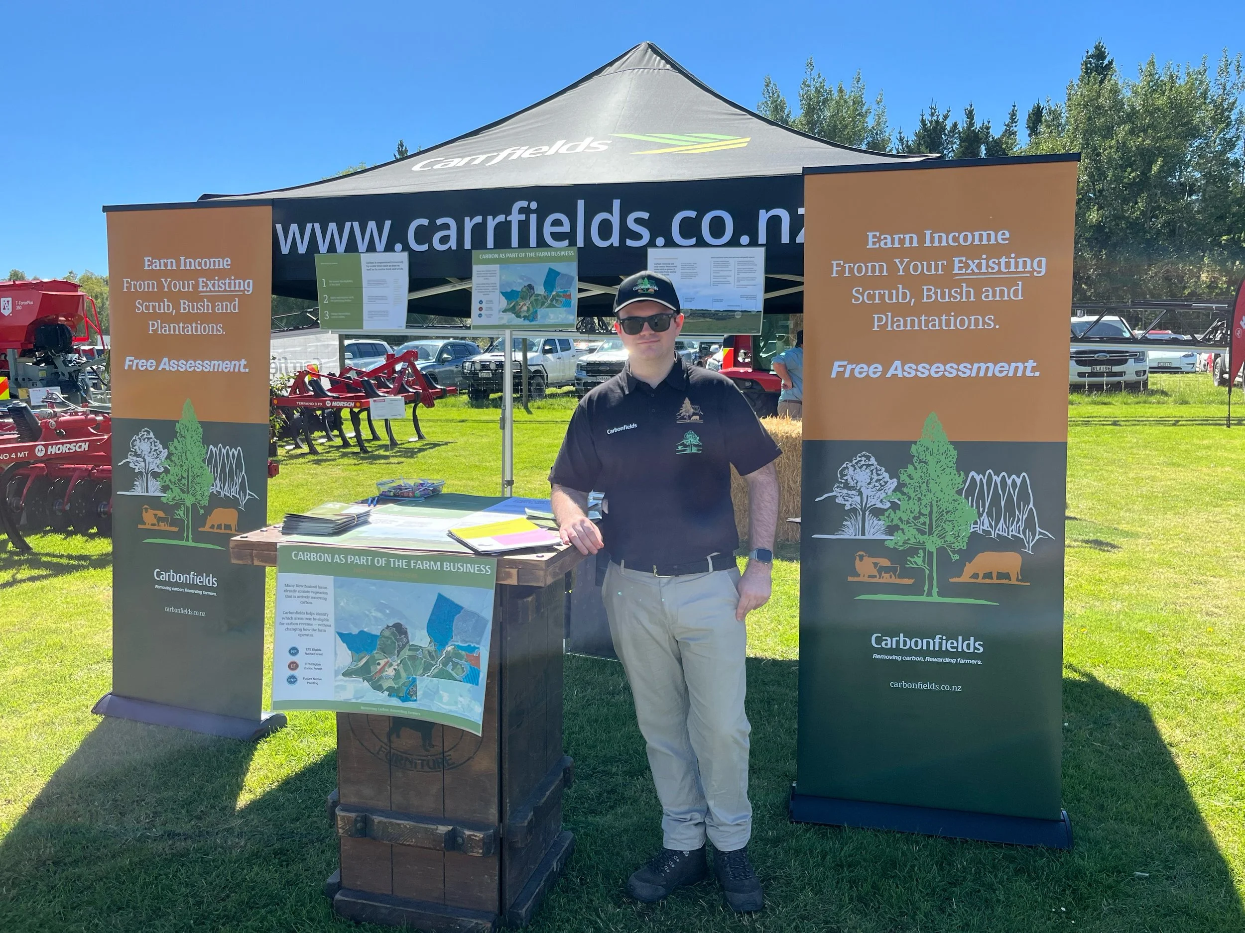 An outdoor booth with a black canopy labeled 'Fields' and a website 'www.carrfields.co.nz'. Two orange banners on either side advertise earning income from existing scrub, bush, and plantations with free assessment. A man wearing a black shirt, white pants, sunglasses, and a cap is standing in front of the booth, which displays informational flyers and maps about carbon farming. The background includes parked cars, trees, and a clear blue sky.