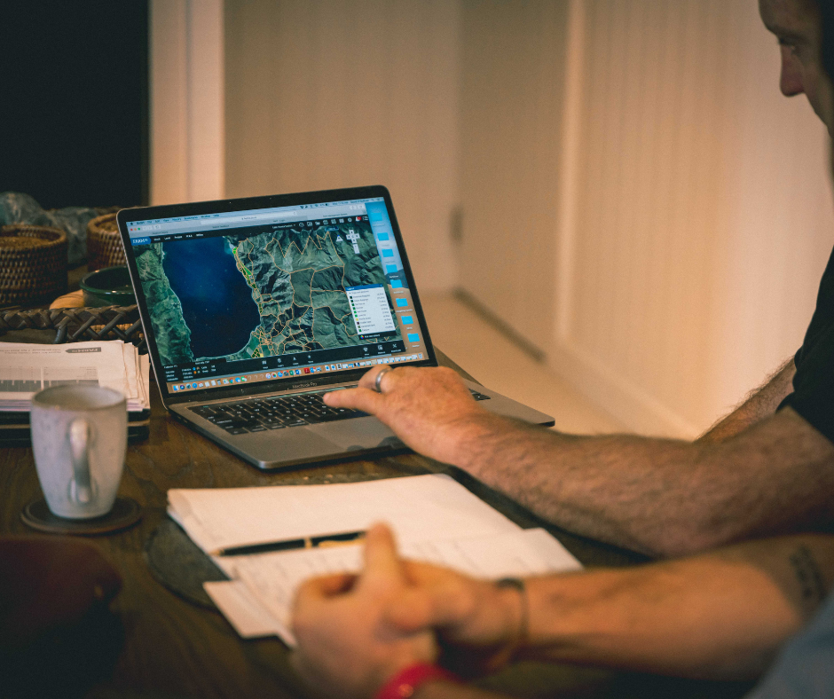 Person working on a laptop with a map displayed on the screen, sitting at a wooden table with papers, a mug, and other items.