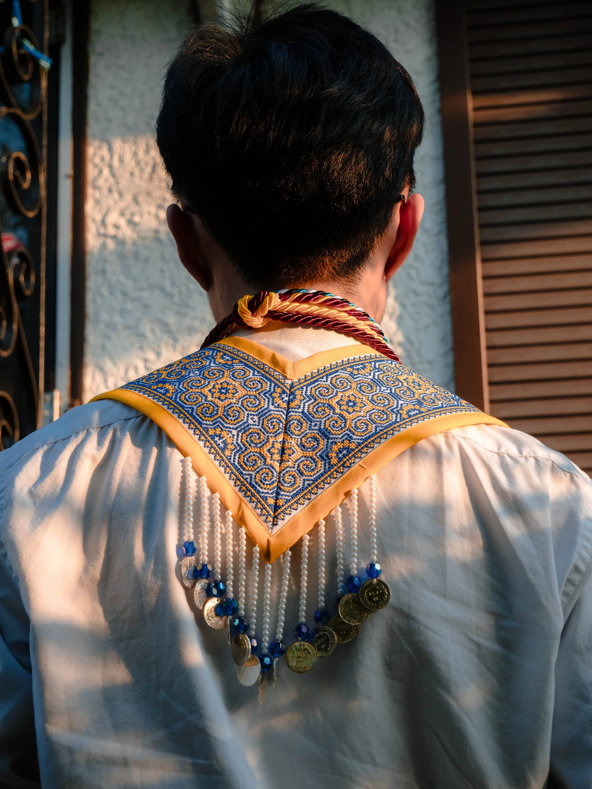 Back of person wearing traditional clothing with a decorated collar and a necklace of coins and beads, standing in front of a textured wall and wooden slats.