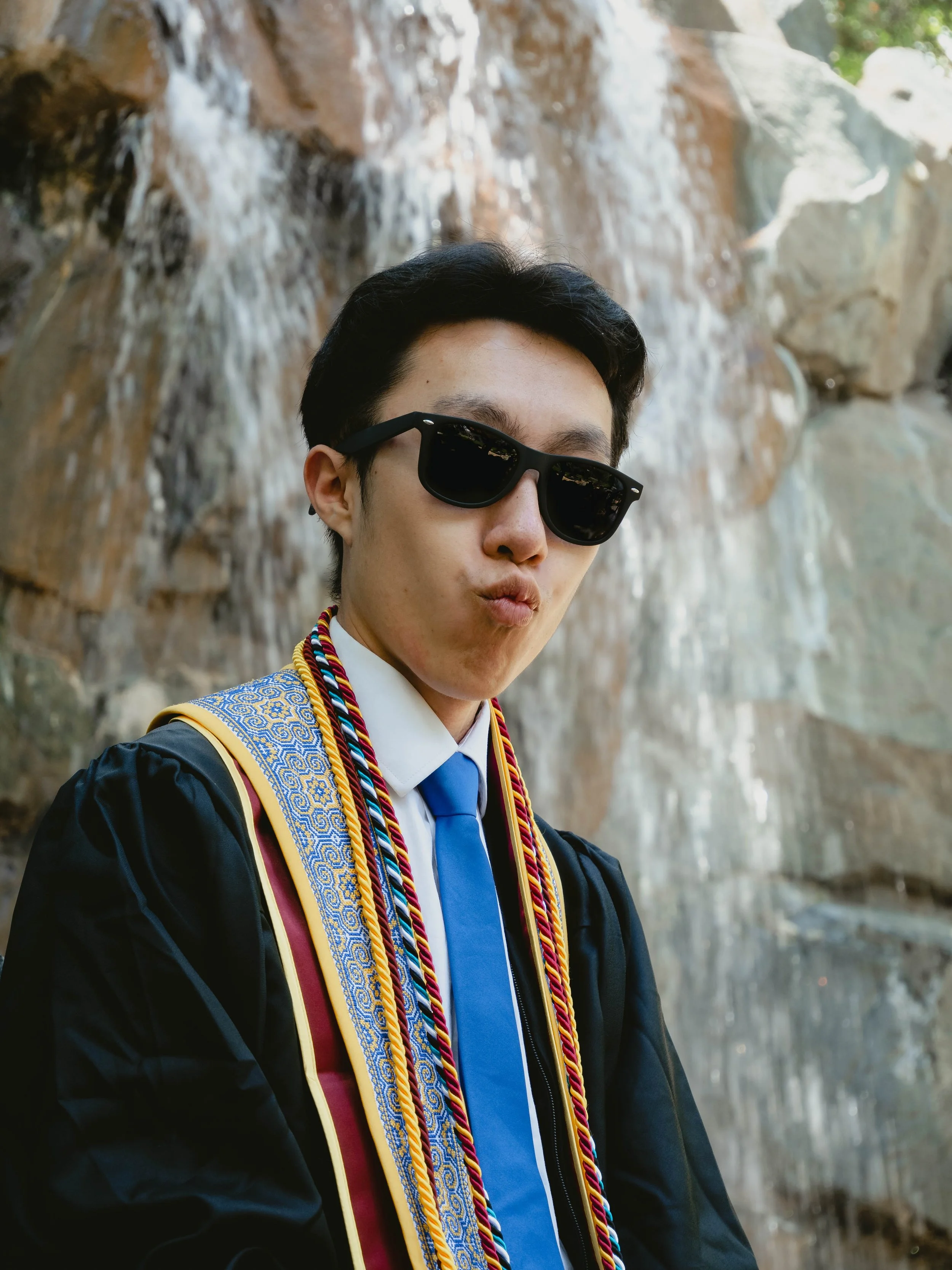 Young man in graduation cap and gown, wearing sunglasses, standing in front of a waterfall.