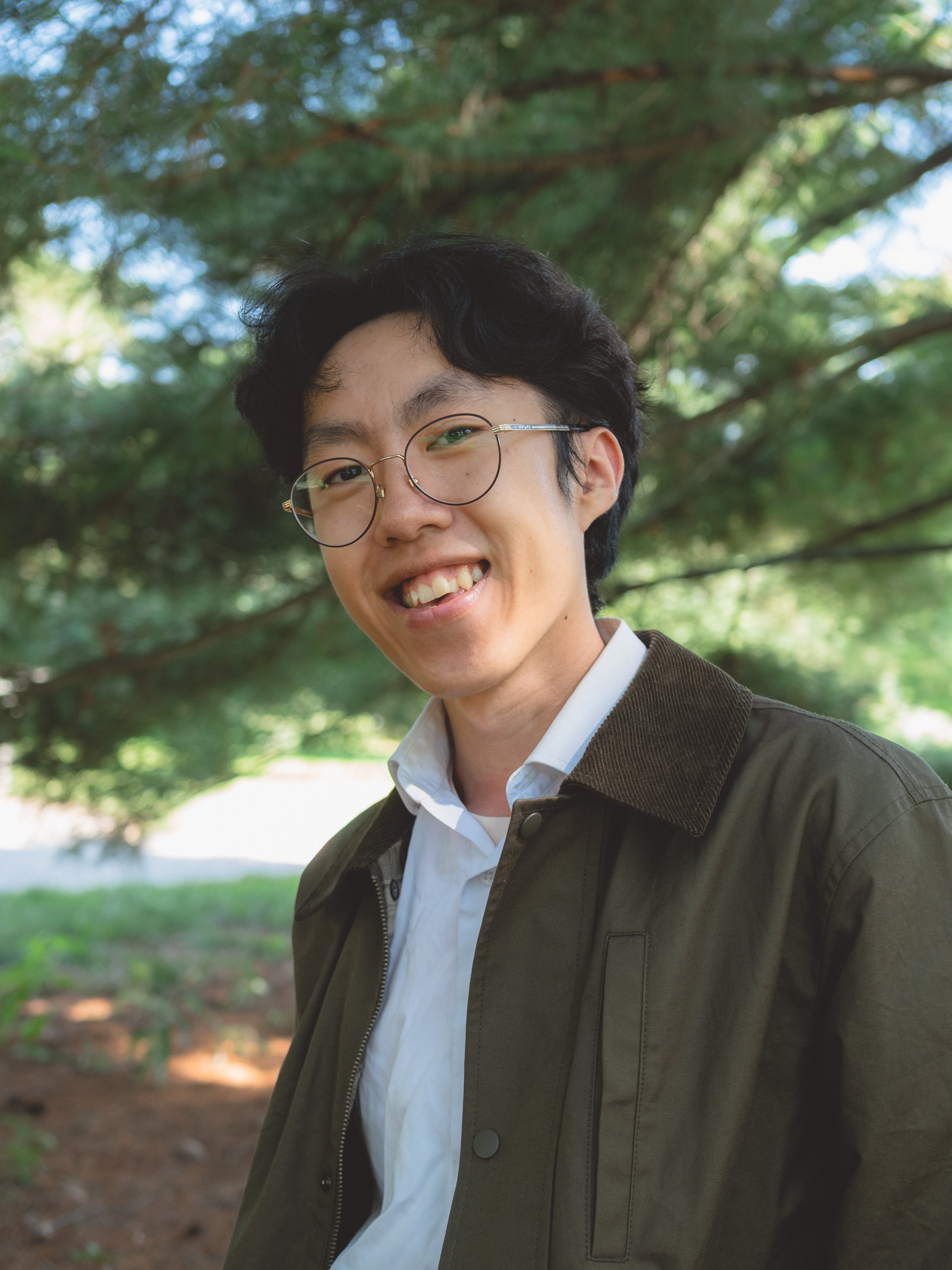 A young man with glasses smiling outdoors in front of green tree foliage.