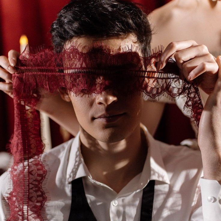 A man holding a red lace mask in front of his eyes, partially obscuring his face, in a dimly lit setting.