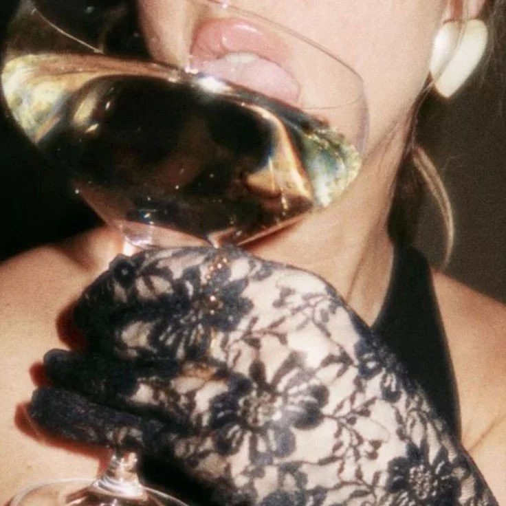 A woman with earrings and a lace-covered hand holding a glass of red wine close to her lips.