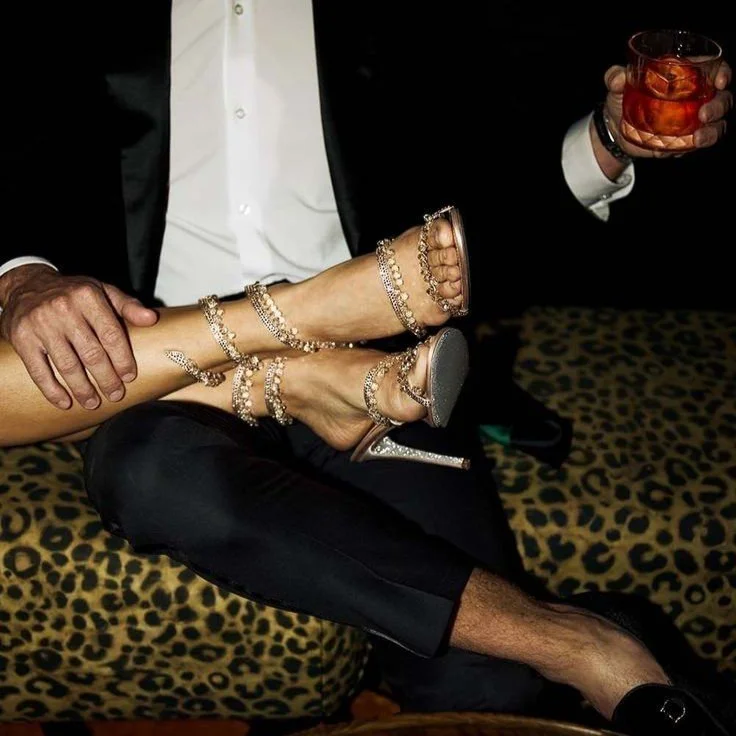 Person in a tuxedo holding a woman's feet, which are in high-heeled sandals with rhinestones, while holding a glass of dark-colored beverage with ice. The scene has a leopard print background.
