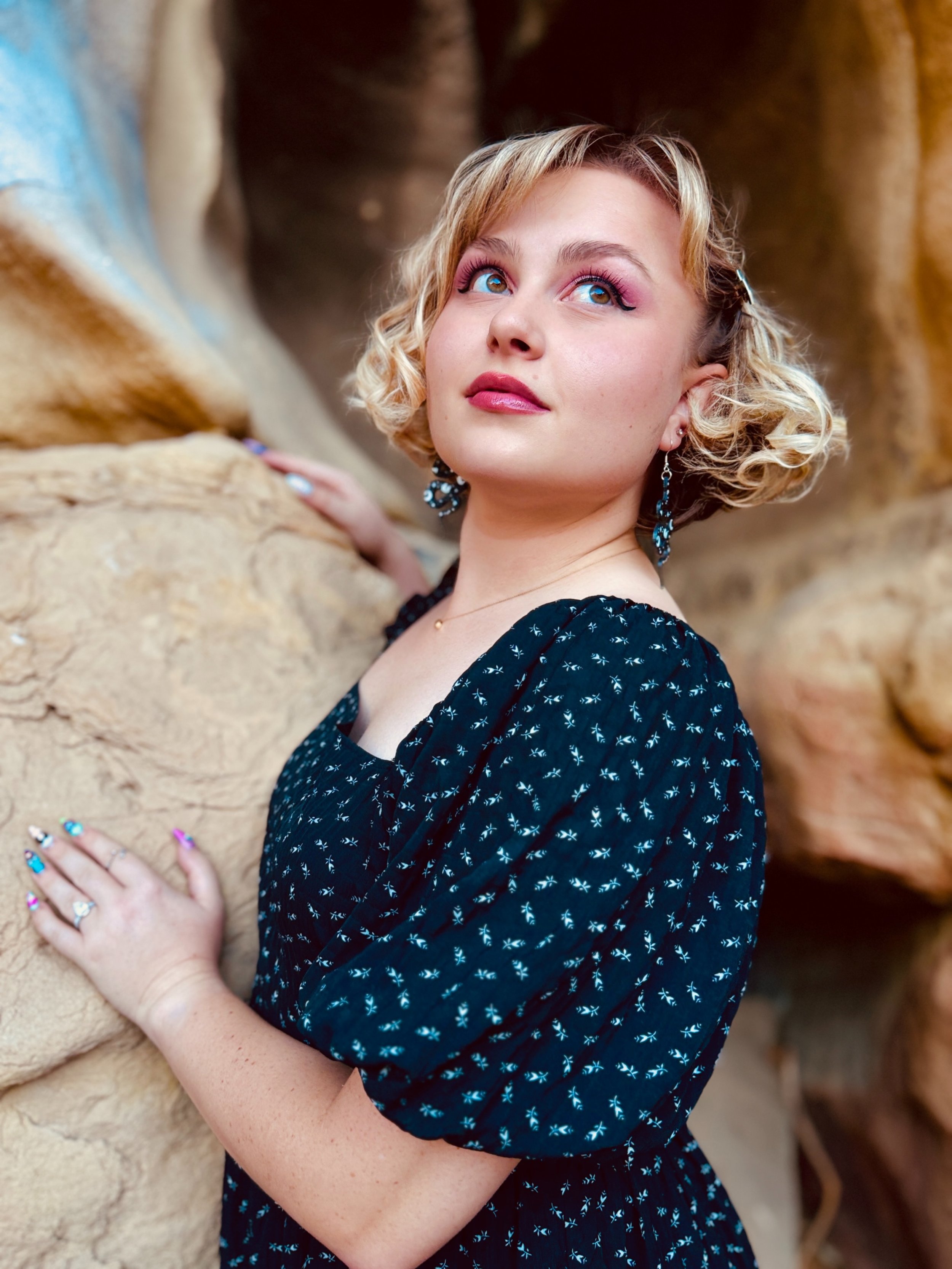 A woman with blonde curly hair and blue eyes wearing a dark blue dress with small white patterns, earrings, and a ring, is touching a beige, textured rock or mountain and looking upwards.