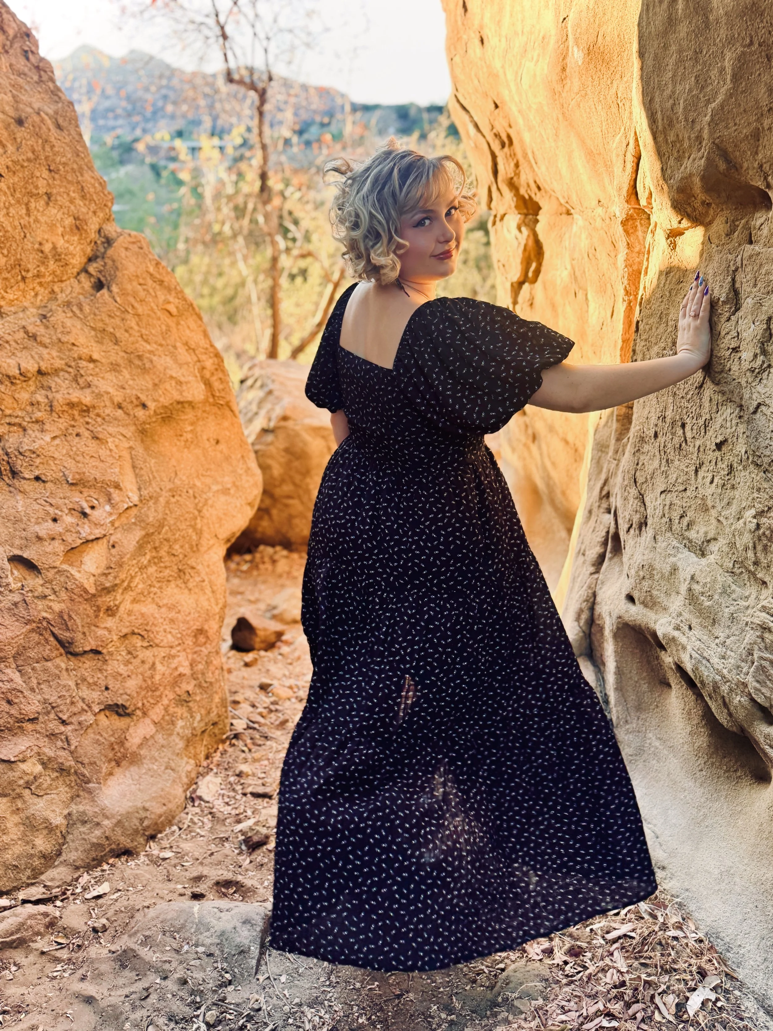 A woman with curly blonde hair and wearing a black dress with white dots stands between large reddish and beige rock formations, touching one of the rocks with her right hand, with trees and a mountain in the background.