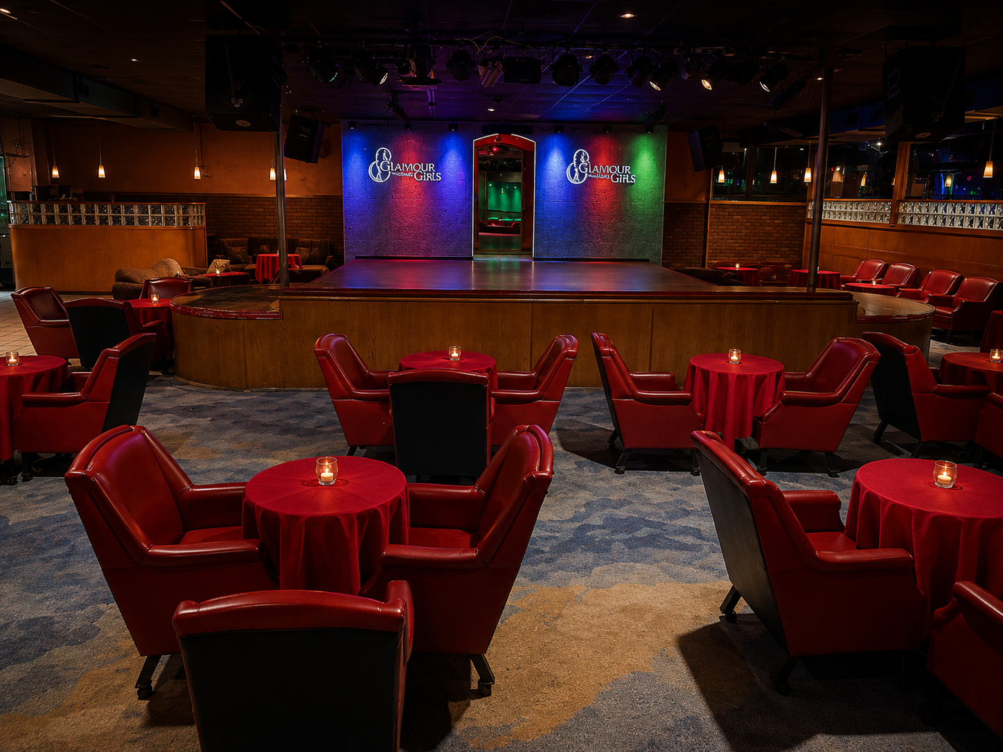 Empty lounge with red chairs and tables, small candles, stage with neon signs saying Glamour Girls, upscale lighting and decor.