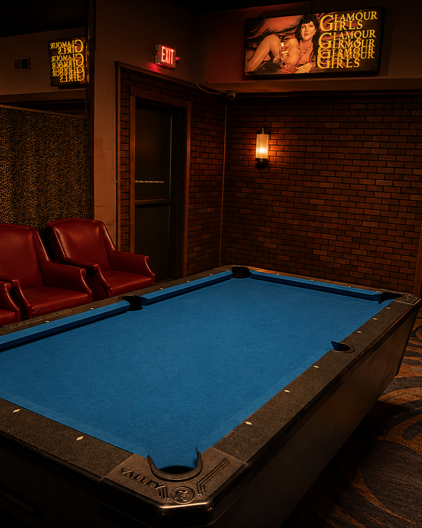 A pool table with a bright blue felt top in a dimly lit room with brick walls, red leather chairs, a wall-mounted lamp, and a television screen displaying an advertisement for Glamour Girls.