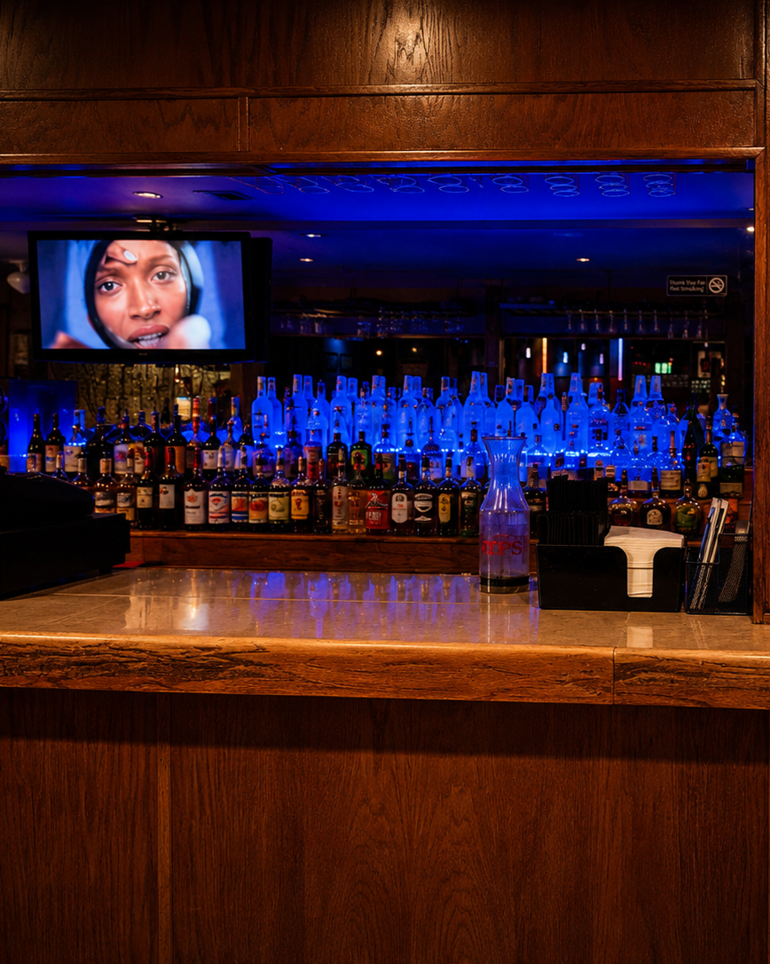 A bar with a variety of liquor bottles illuminated by blue backlighting, a TV screen showing a close-up of a woman's face, and a wooden counter with a plastic pitcher, napkins, and a container for straws or stir sticks.