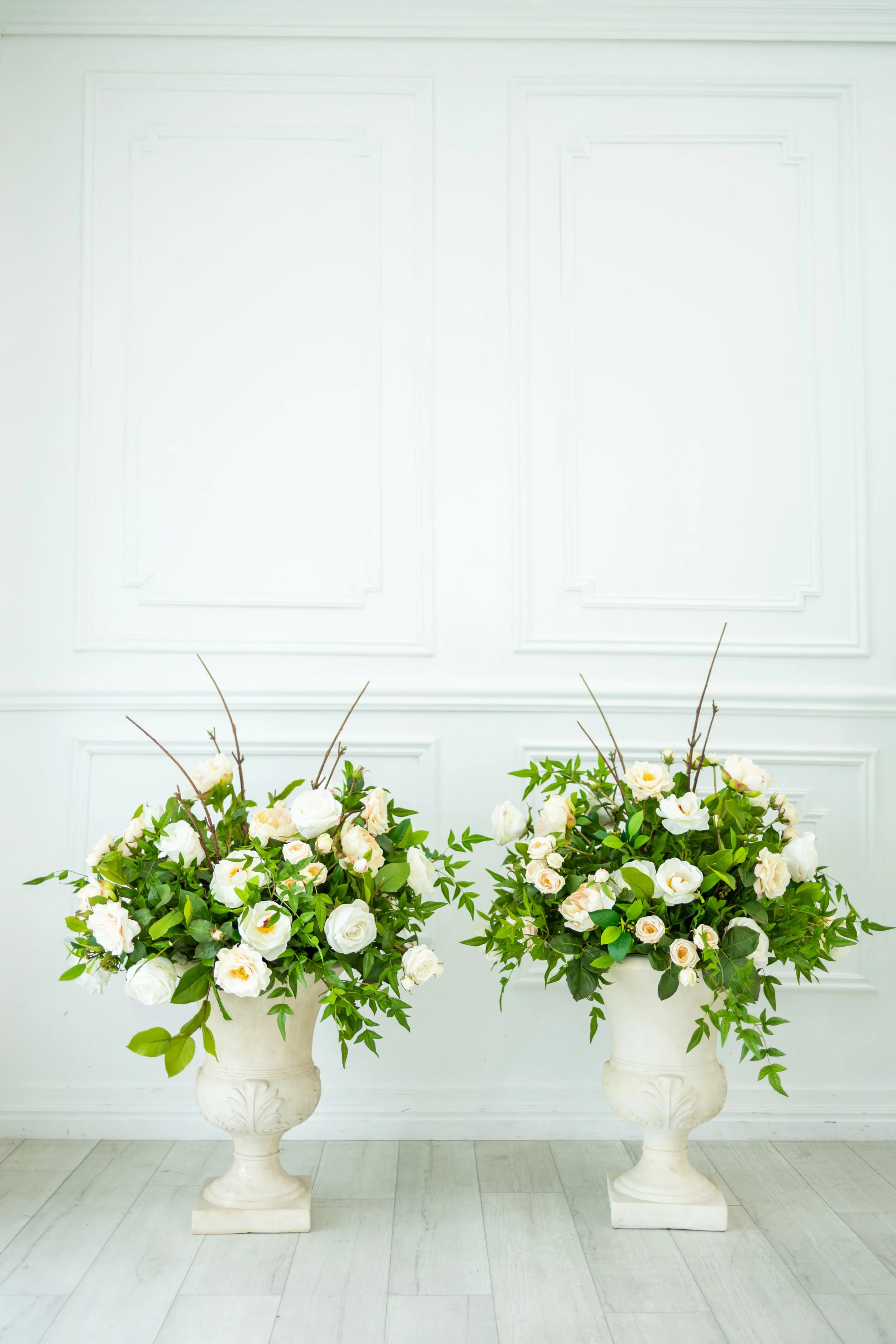 Two large floral arrangements in white vases with white and light pink flowers and green foliage, set against a white wall with decorative paneling.