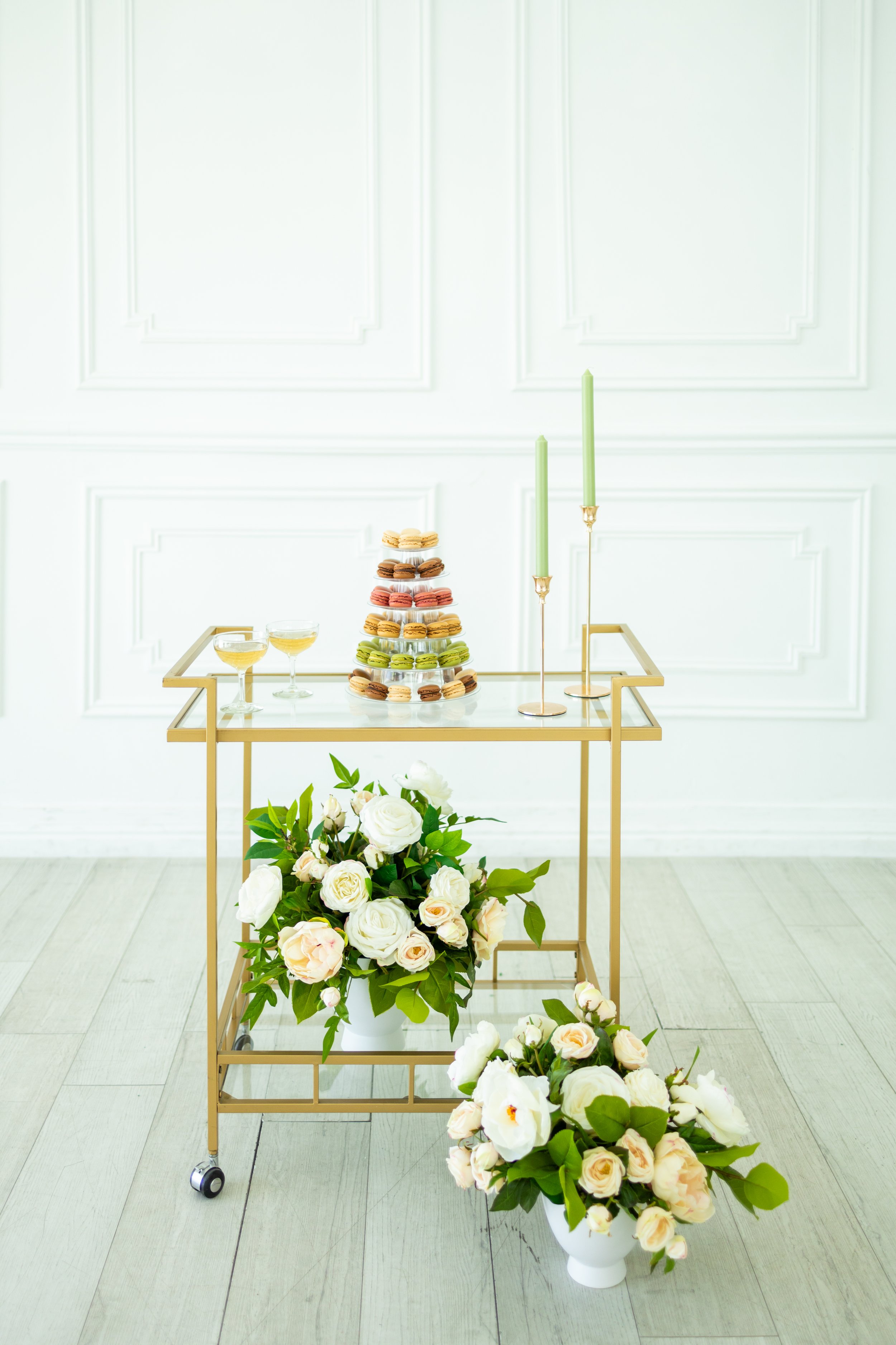 A gold serving cart with a tiered display of colorful macarons, two green candles, and two champagne glasses, with a large bouquet of white and pale pink roses in front, against a white paneled wall.