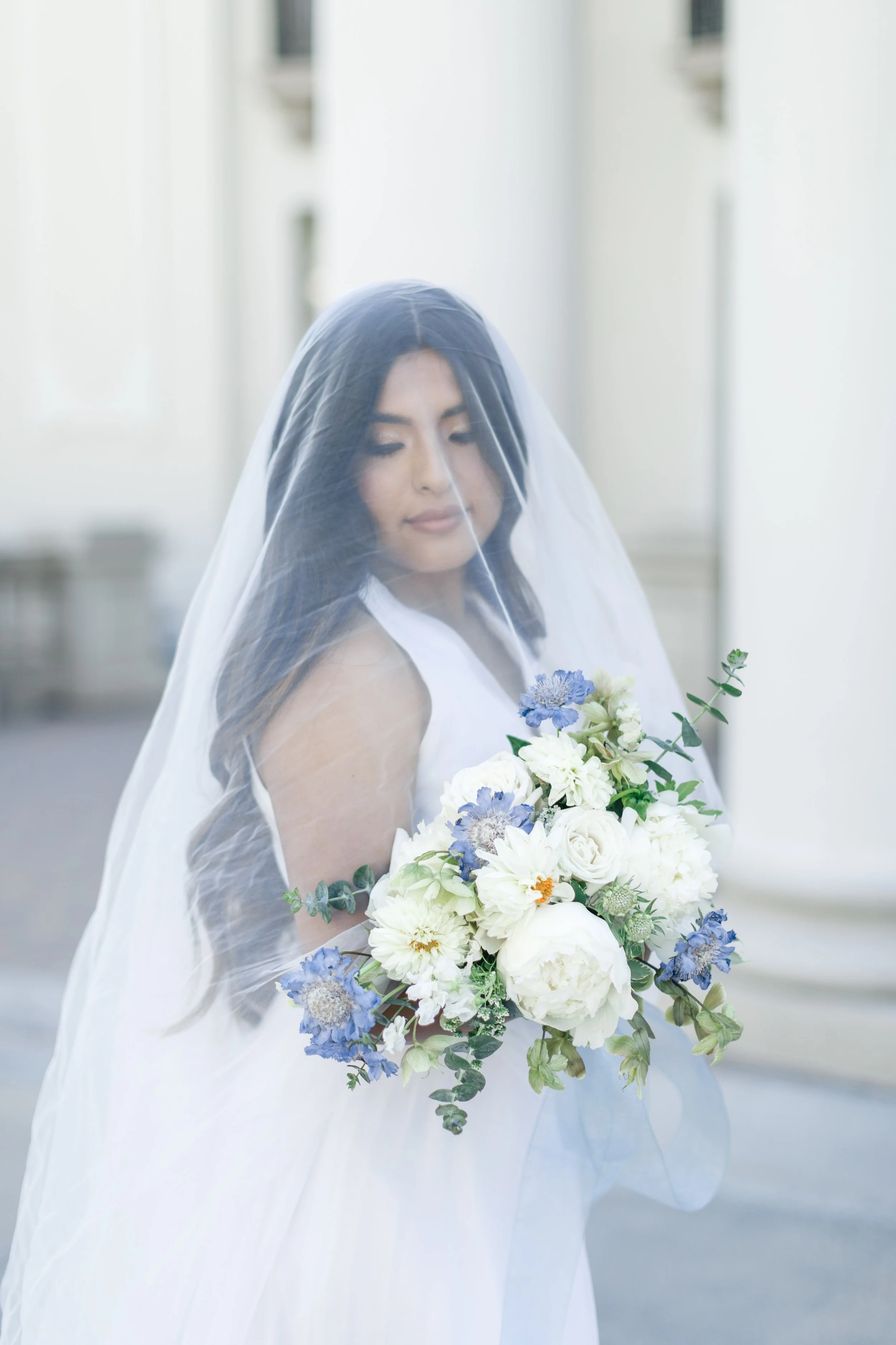 A woman in a white dress holding a bouquet of white and blue flowers, with a white veil on her head, standing outdoors with blurred architectural background.