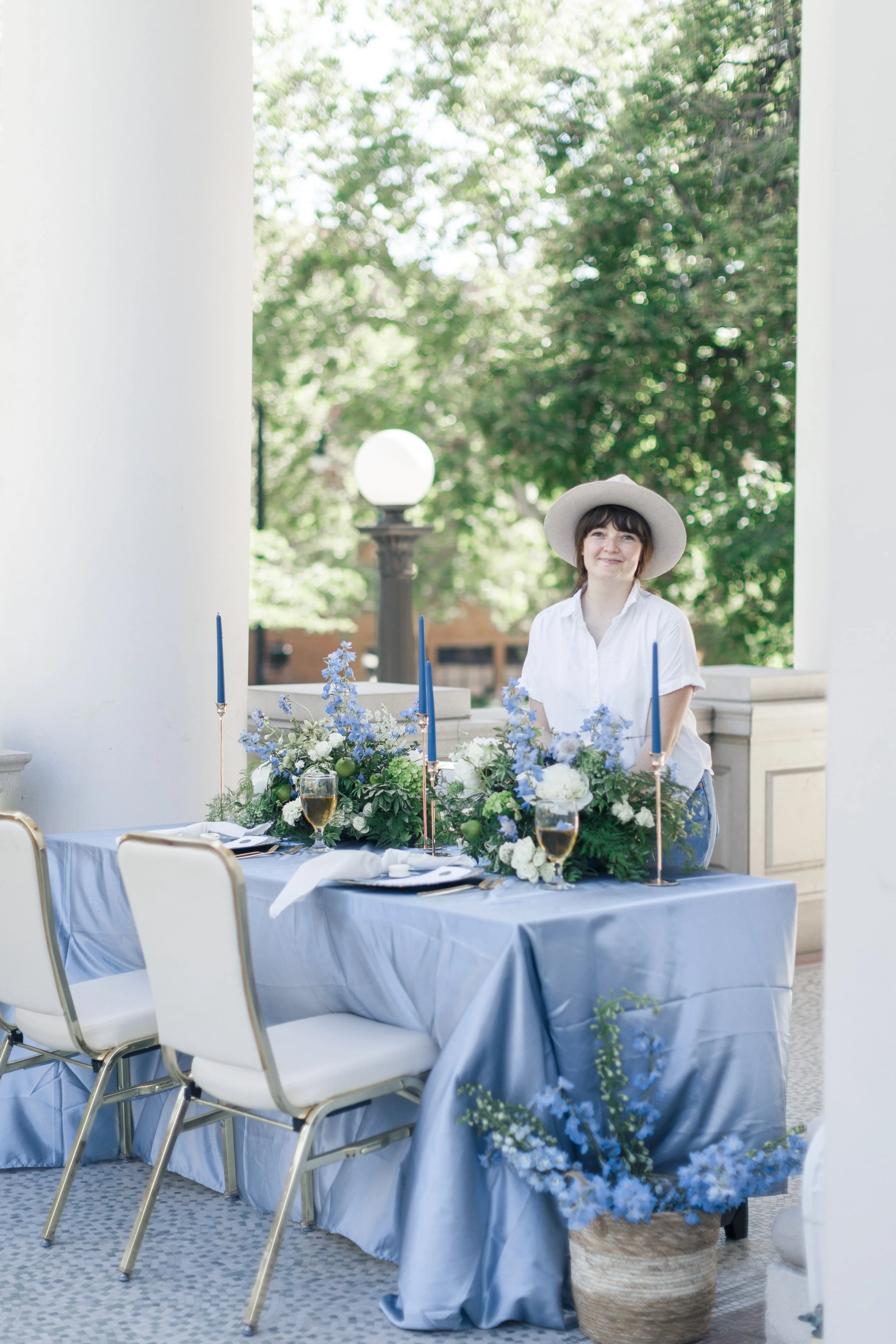 A woman with short dark hair, wearing a wide-brimmed hat and white shirt, standing behind a long outdoor table decorated with blue and white floral arrangements, candlesticks, and glasses of champagne, in a garden setting with trees and a lamp post in the background.