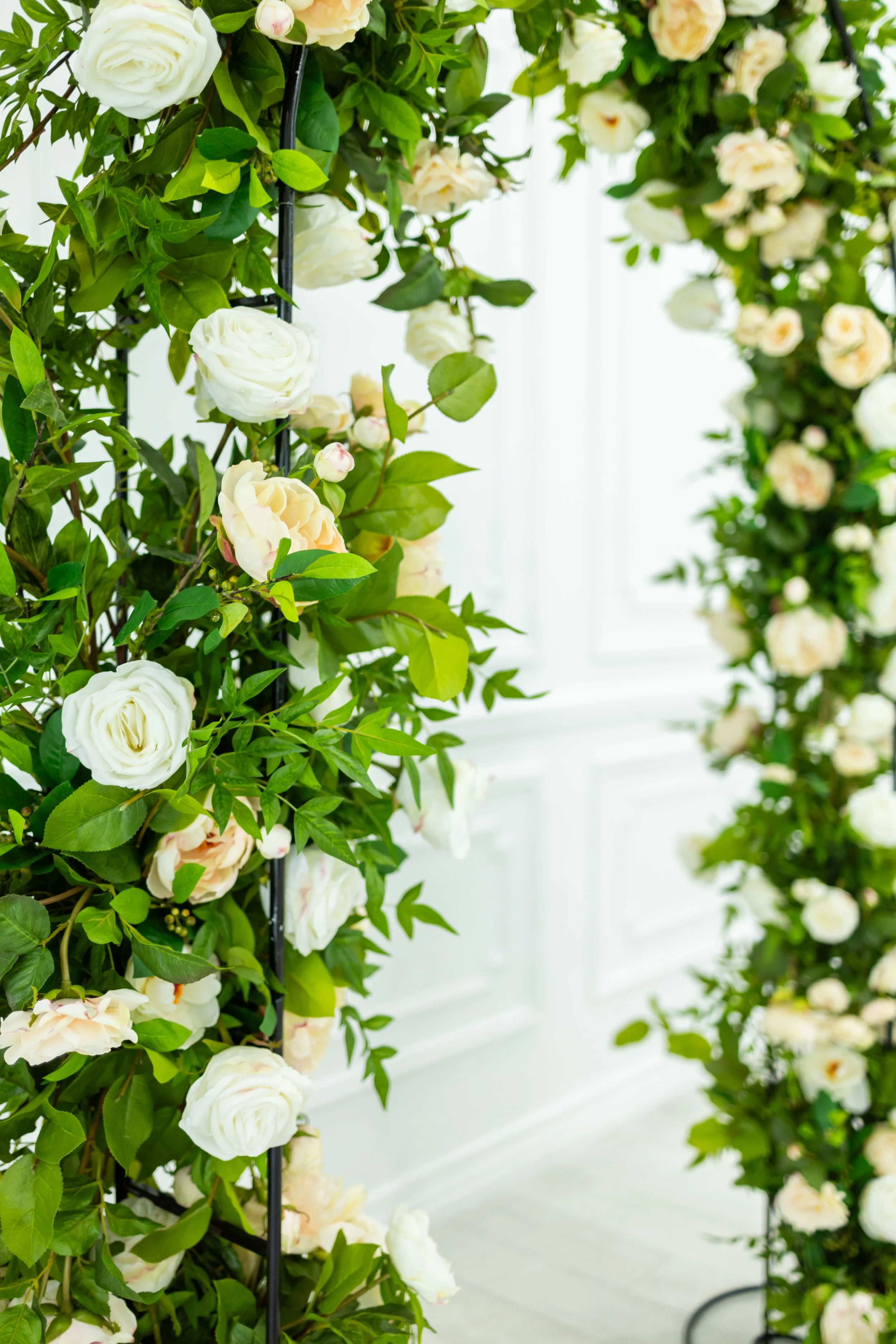 White and cream roses with green leaves arranged on black metal stands in a bright, white room.