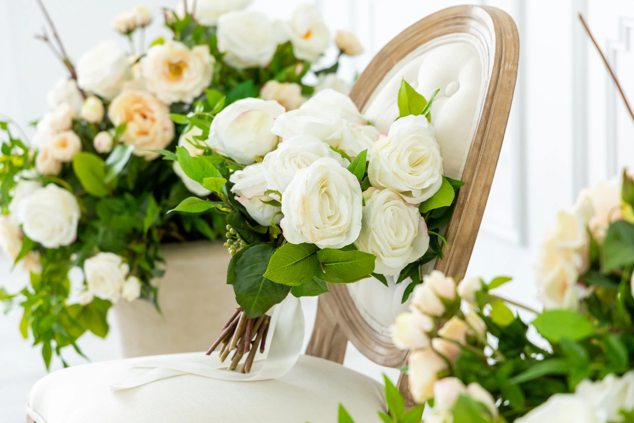A white decorative chair with a bouquet of white roses and green leaves resting on it, surrounded by additional floral arrangements.