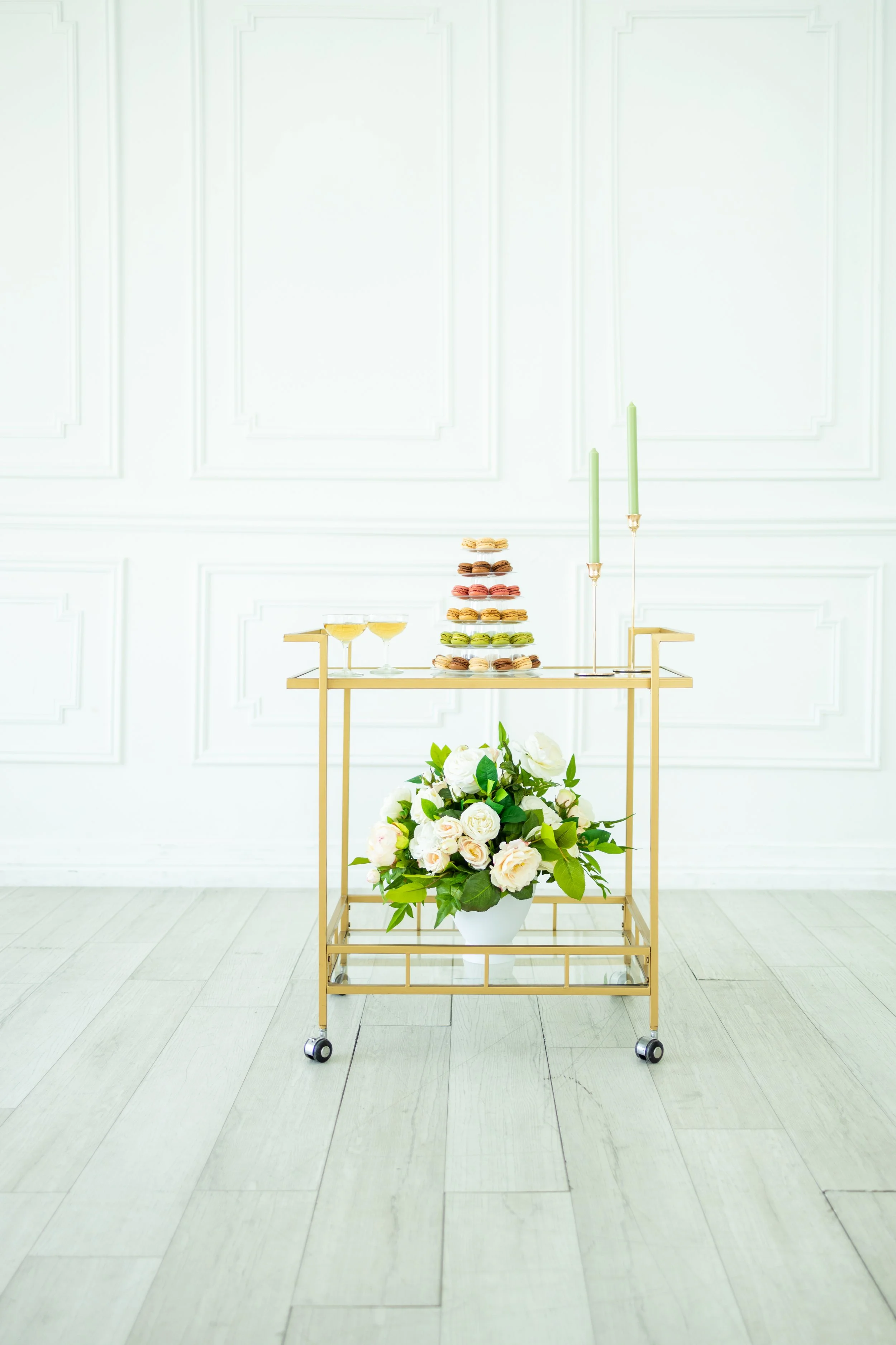 A gold serving cart with four wheels, decorated with flowers and macarons, set against a white paneled wall with a minimalist, elegant décor.