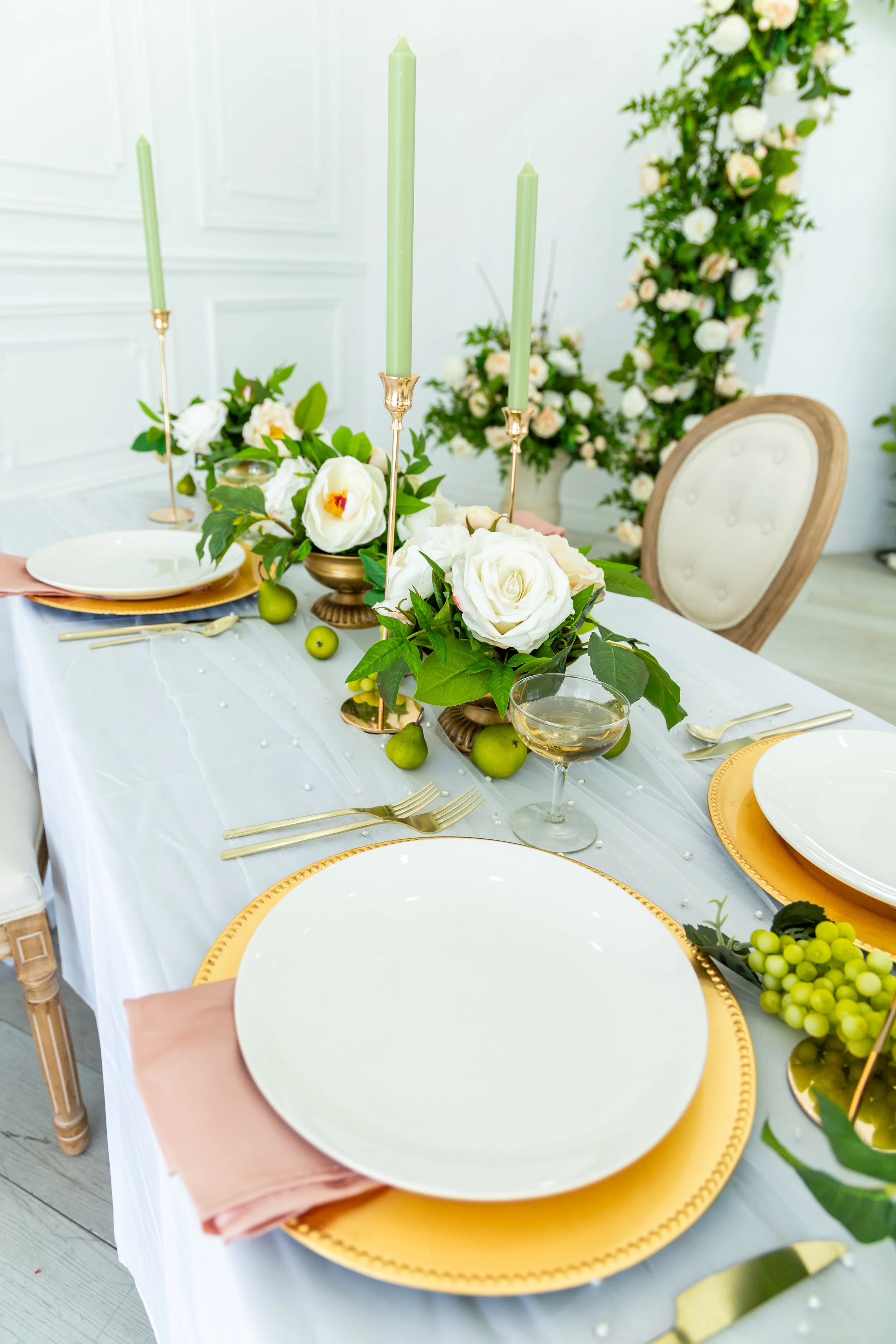 Elegant dining table setup with white plates, gold-rimmed charger plates, and blush pink napkins, decorated with white roses, greenery, tall candles in gold holders, green grapes, and glassware, all set against a backdrop of white walls and floral ar